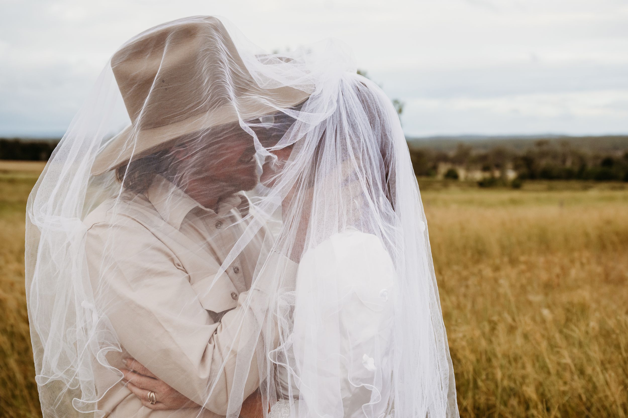 Bride and groom sharing a romantic kiss under a wedding veil in a golden field.