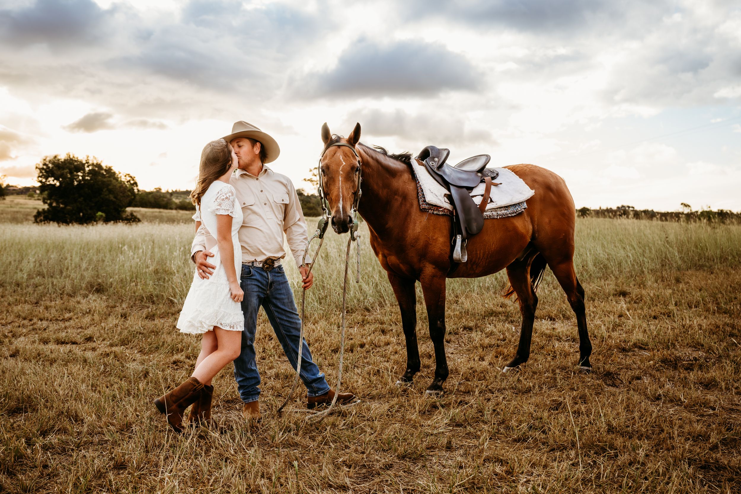 A couple in western attire walks with a saddled brown horse through a golden field at sunset.
