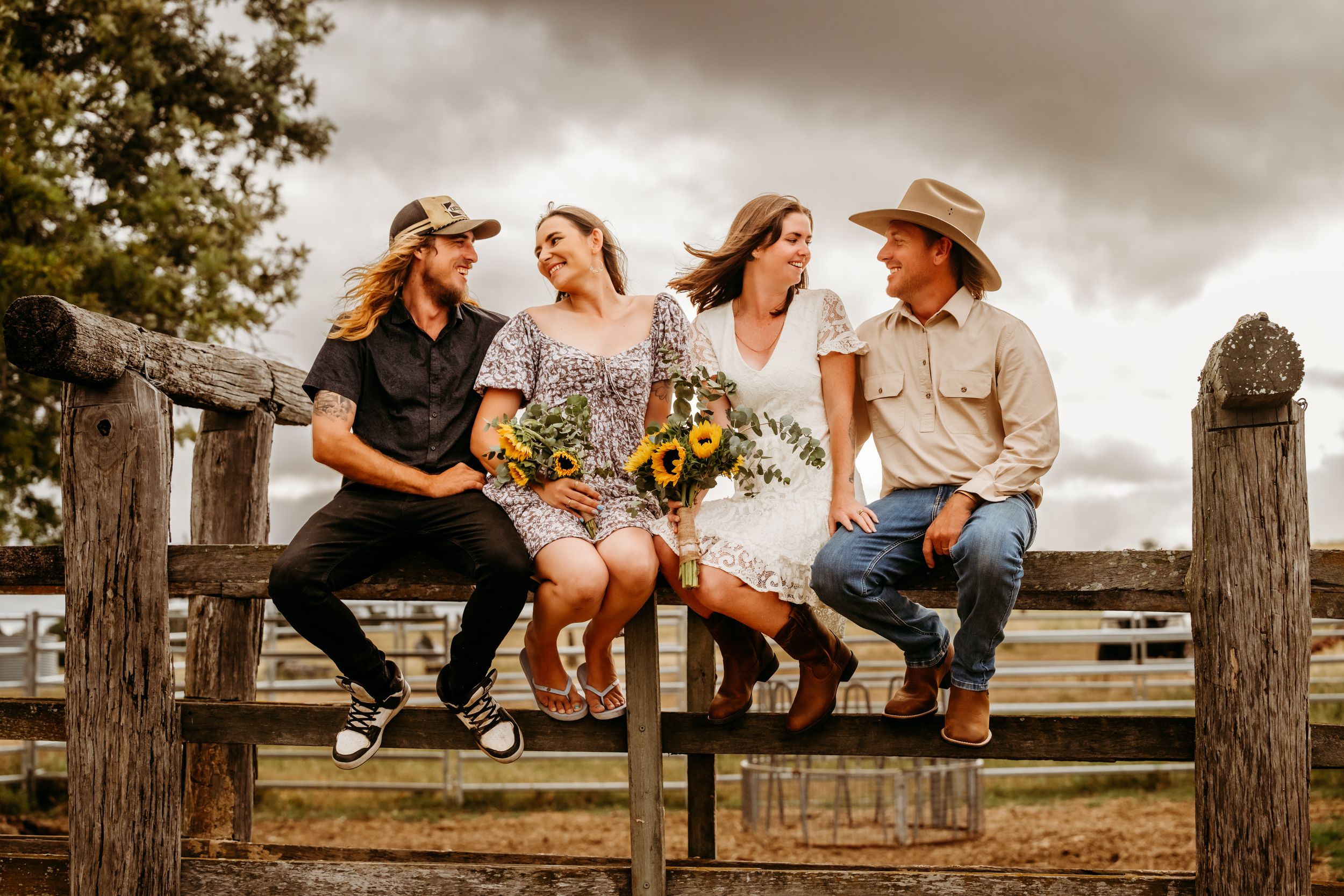 Four friends sit on a rustic wooden fence laughing with sunflowers on a cloudy day.
