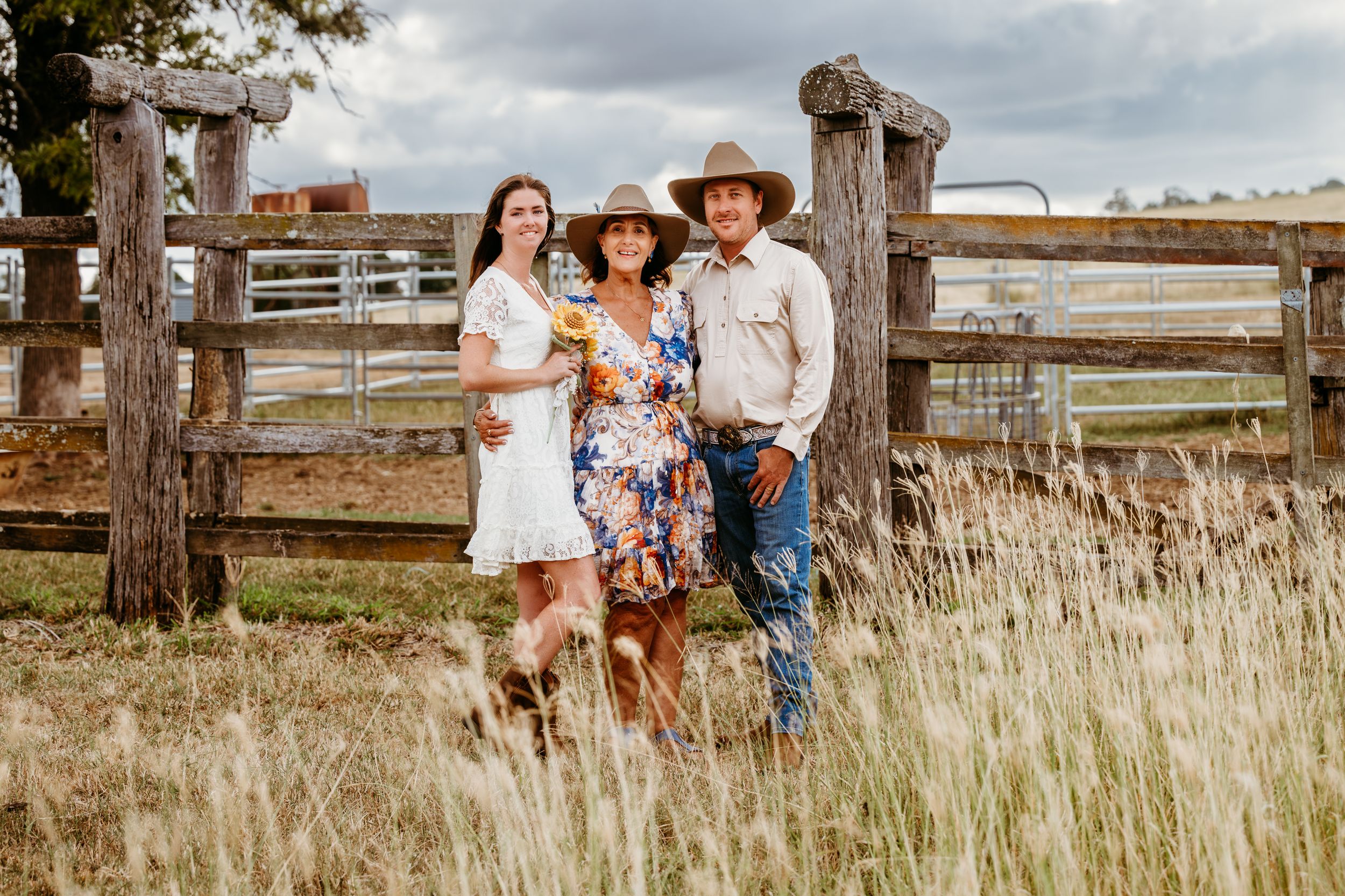 Three people in western attire pose by a rustic wooden fence in a grassy field.
