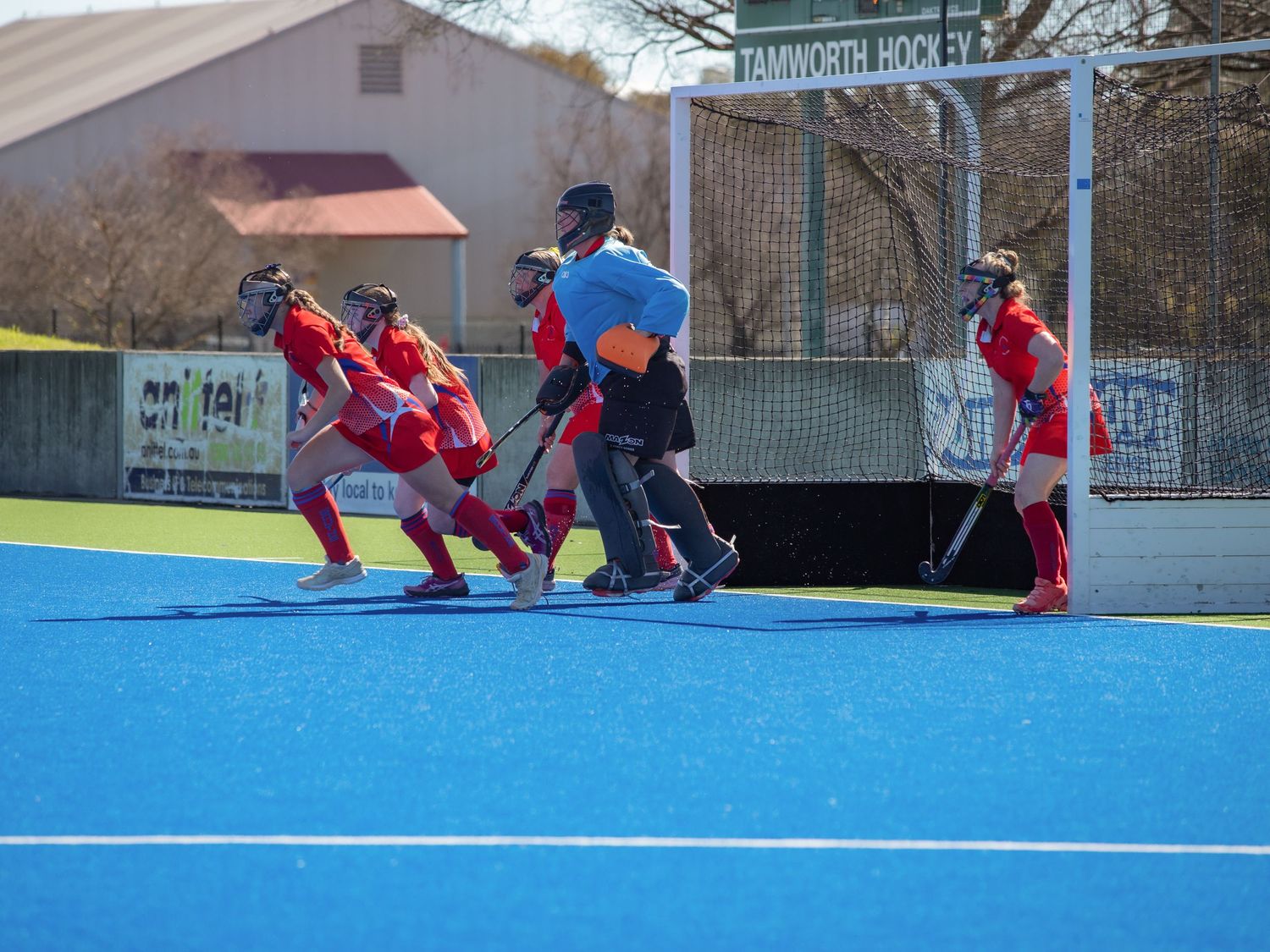 Field hockey players in red uniforms compete on a blue turf pitch at Tamworth Hockey Club on a sunny day.
