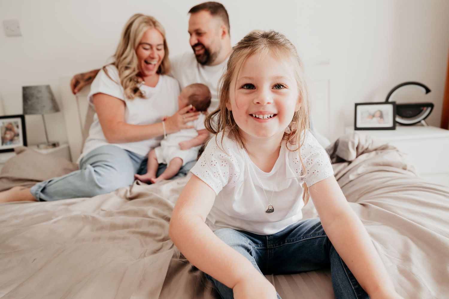 Young girl smiling on bed with parents and newborn baby in bright bedroom family lifestyle photo.