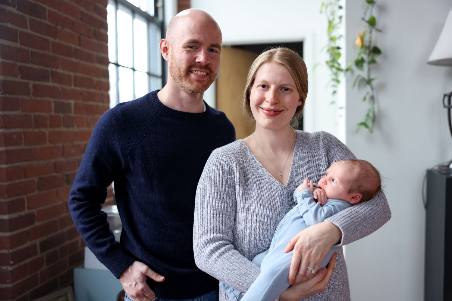 Smiling new parents pose indoors holding their newborn baby wrapped in a light blue blanket.