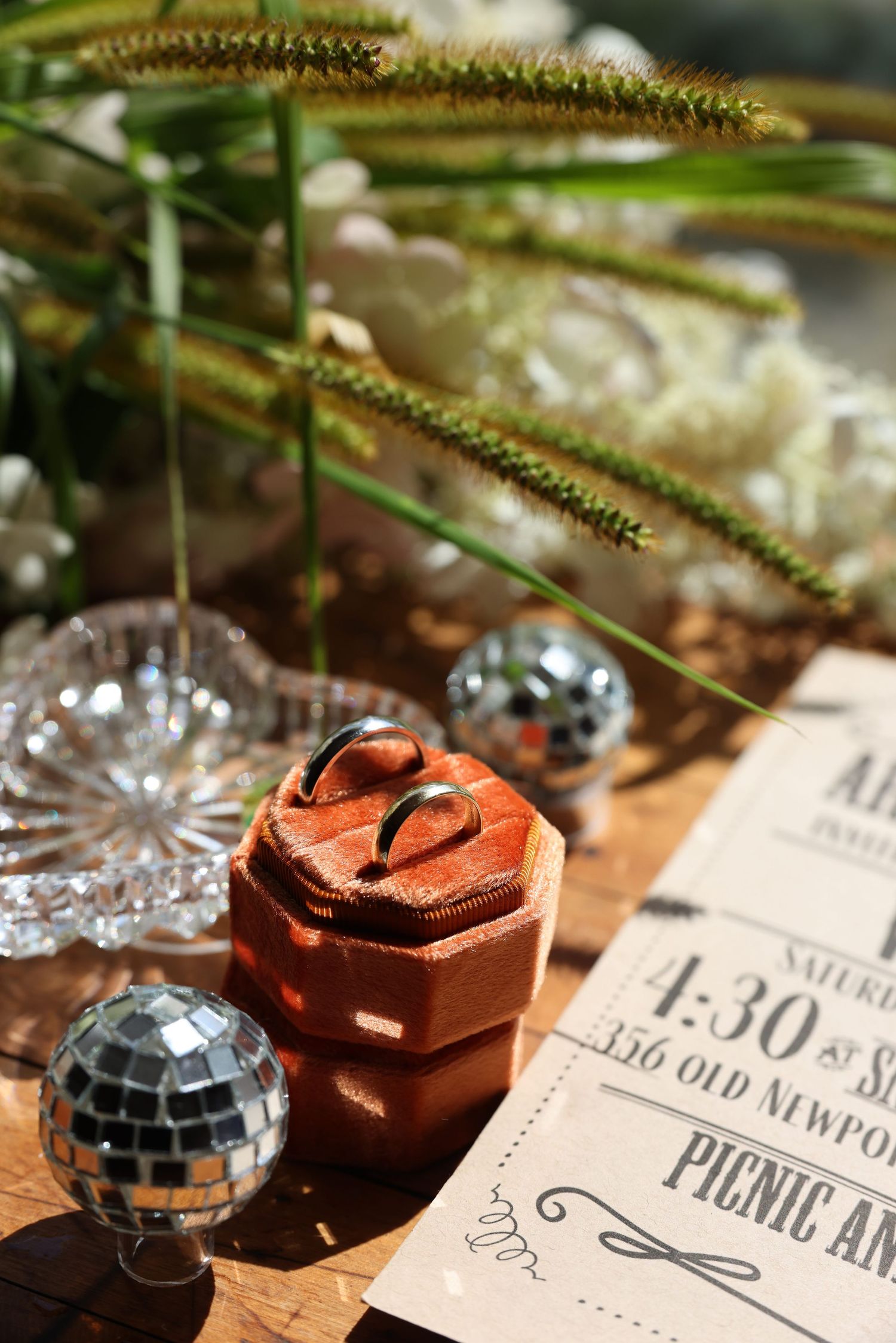 Wedding ring in an orange velvet box surrounded by mini disco balls and invitation on a wooden surface.