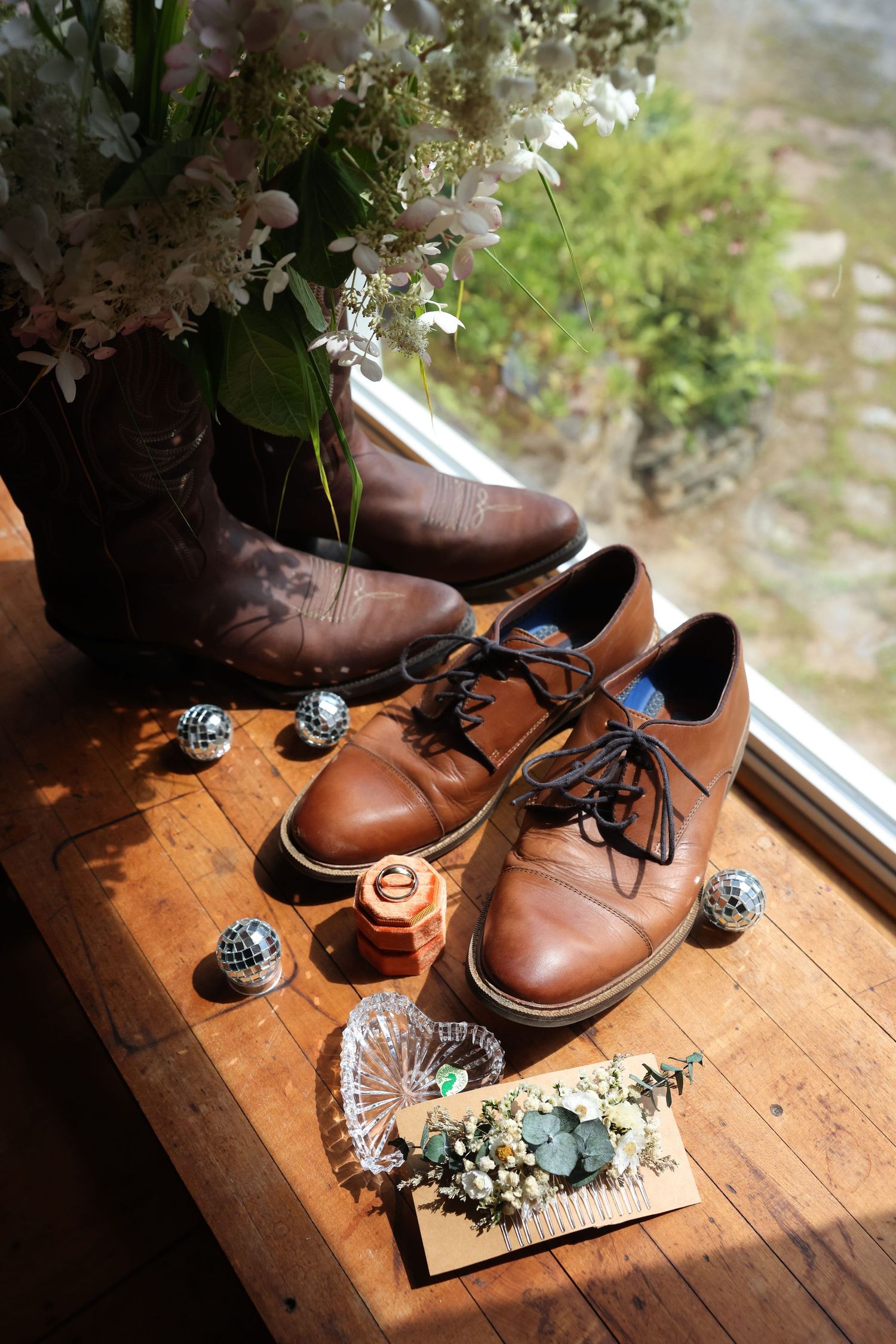Brown cowboy boots and leather dress shoes displayed on wooden surface with disco balls and dried floral bouquet.