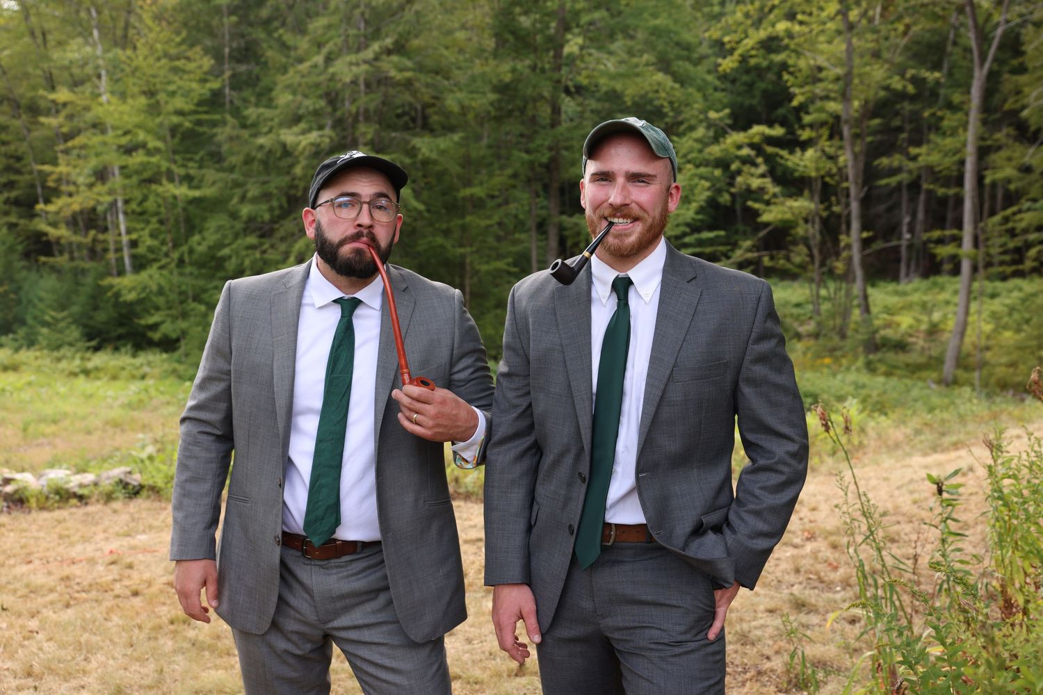 Two men in grey suits with green ties standing outdoors in a wooded field setting.