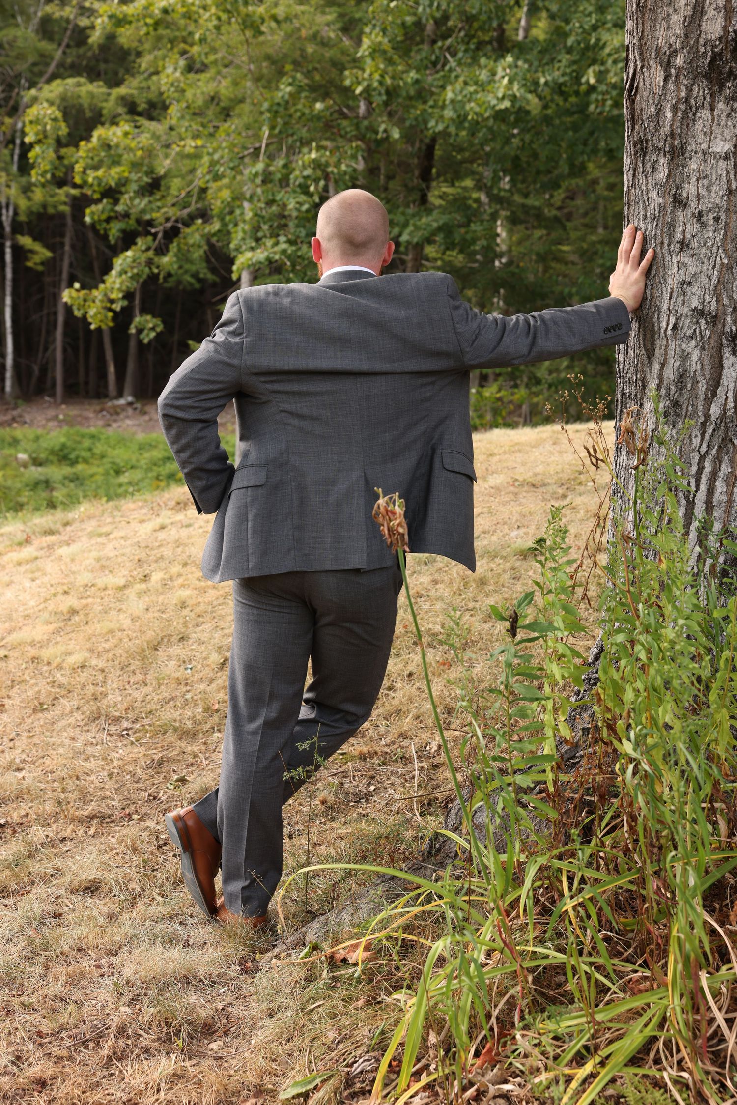 Bald groomsman in gray suit leaning against a tree in an outdoor field viewed from behind.