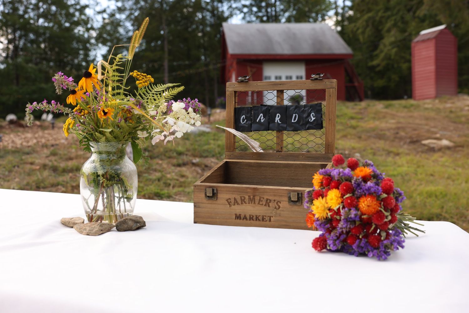 Rustic wedding card box labeled Farmers Market with wildflower arrangements on a white table outdoors.