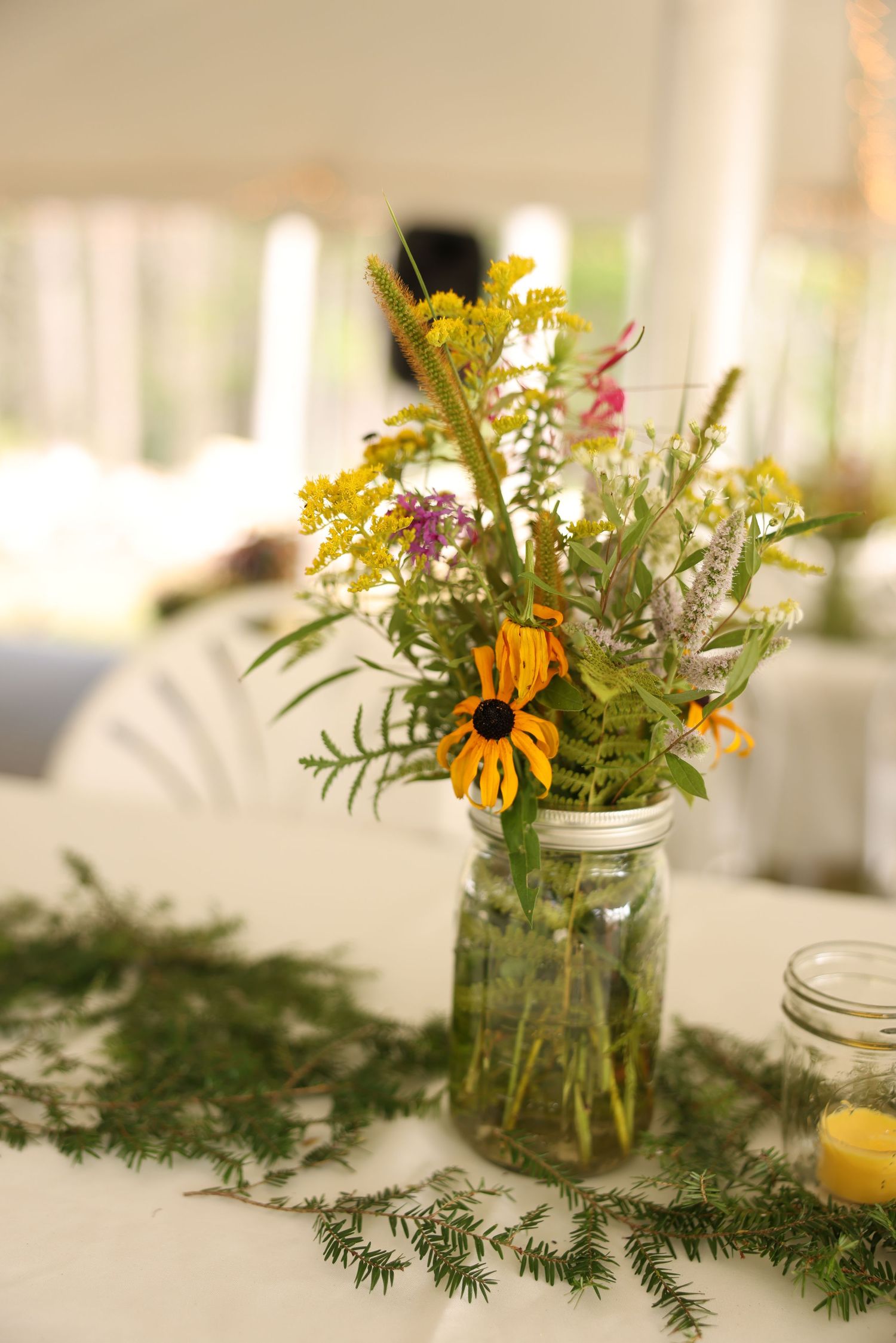 Rustic wildflower centerpiece in mason jar with black-eyed Susans on white tablecloth at outdoor wedding reception.