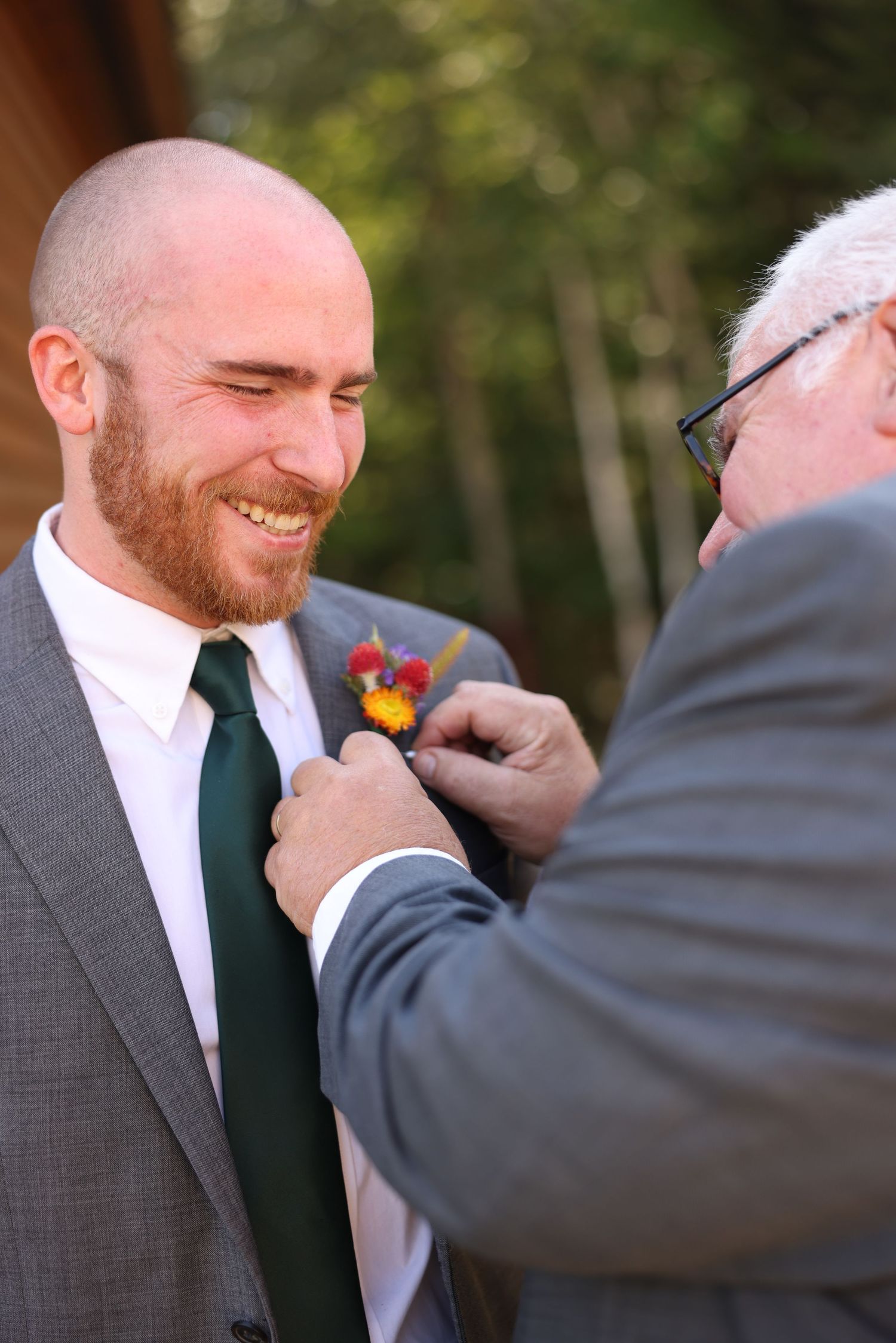 Smiling bald groom with red beard in gray suit gets colorful boutonniere pinned by older man outdoors.