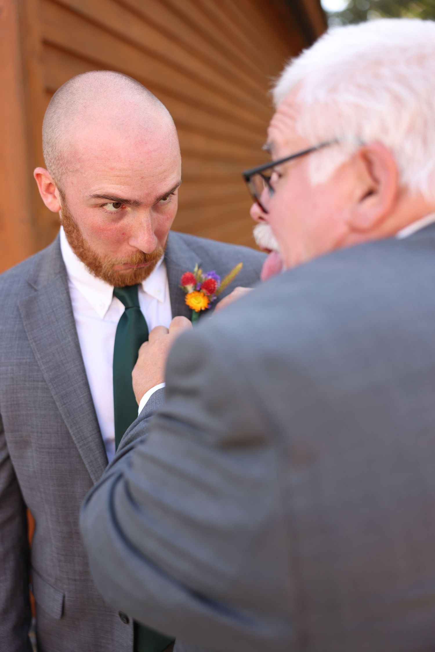 Bald groom with red beard in gray suit and green tie has boutonniere adjusted by older man in glasses.