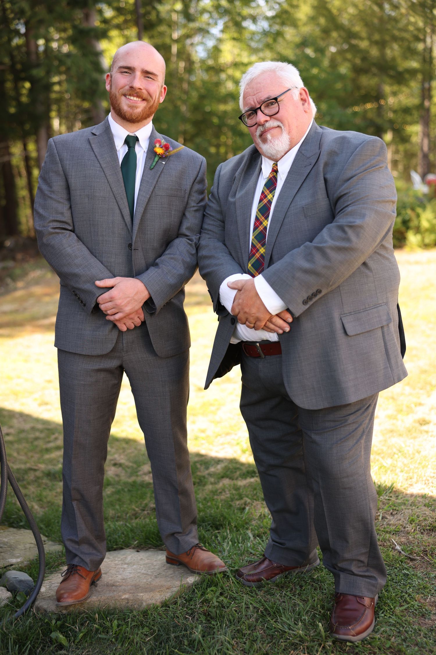 Groom in gray suit with green tie and boutonniere poses with older man in plaid tie at outdoor wedding.