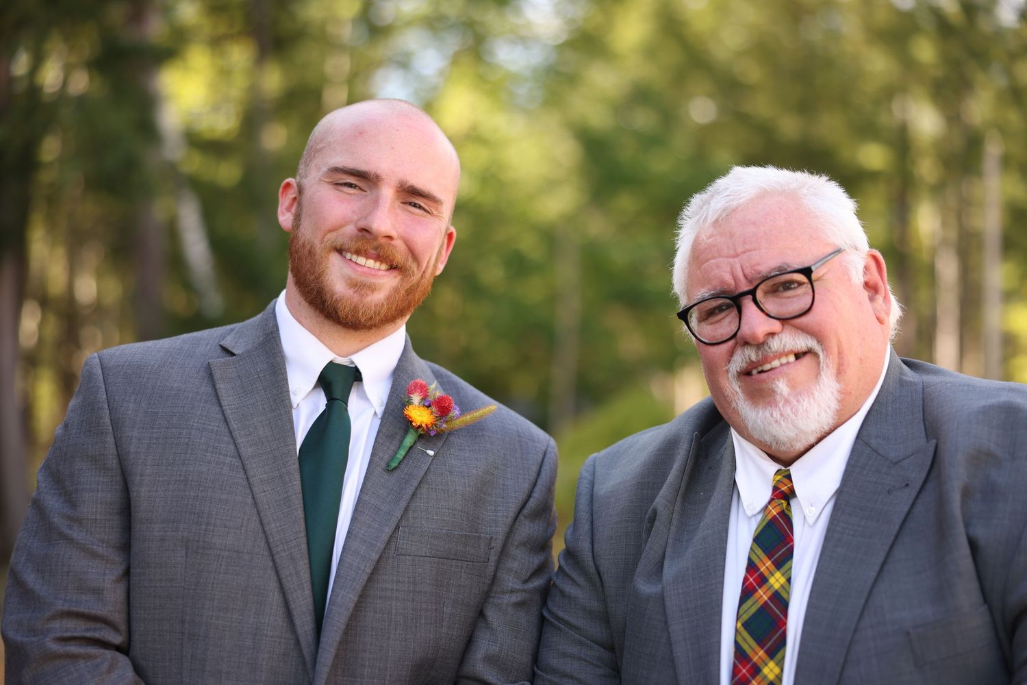Smiling groom in gray suit with colorful boutonniere stands beside older man in plaid tie outdoors.