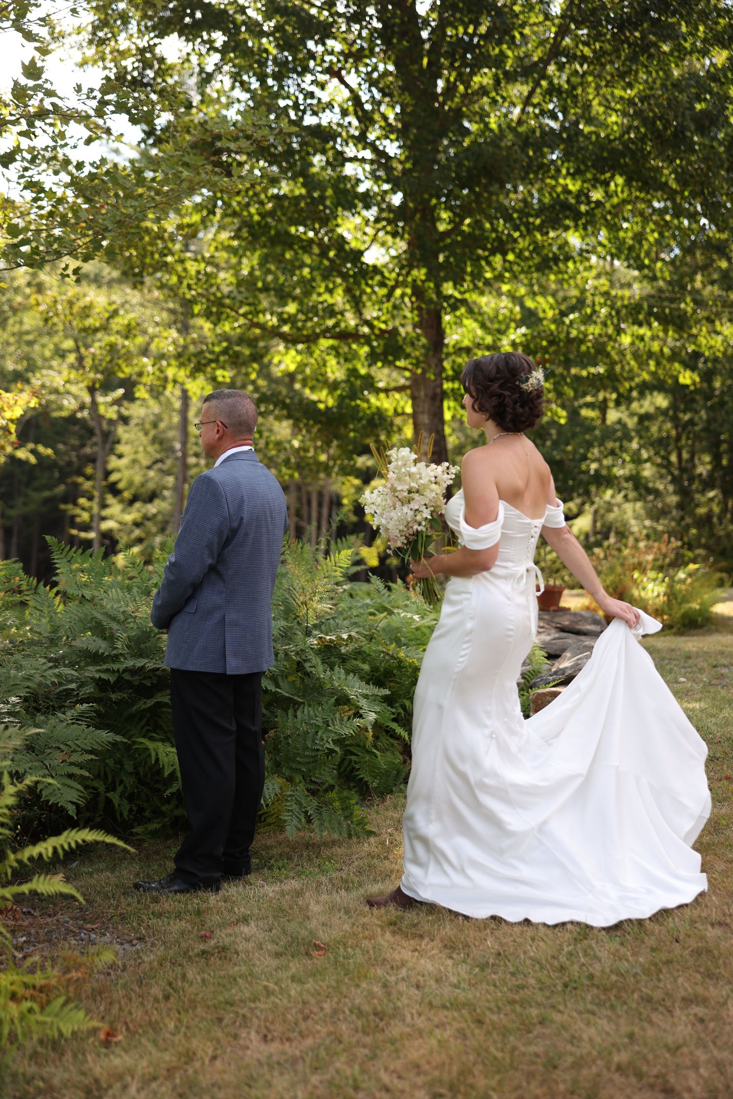 Bride in off-shoulder white gown holding bouquet approaches groom from behind in lush garden setting.