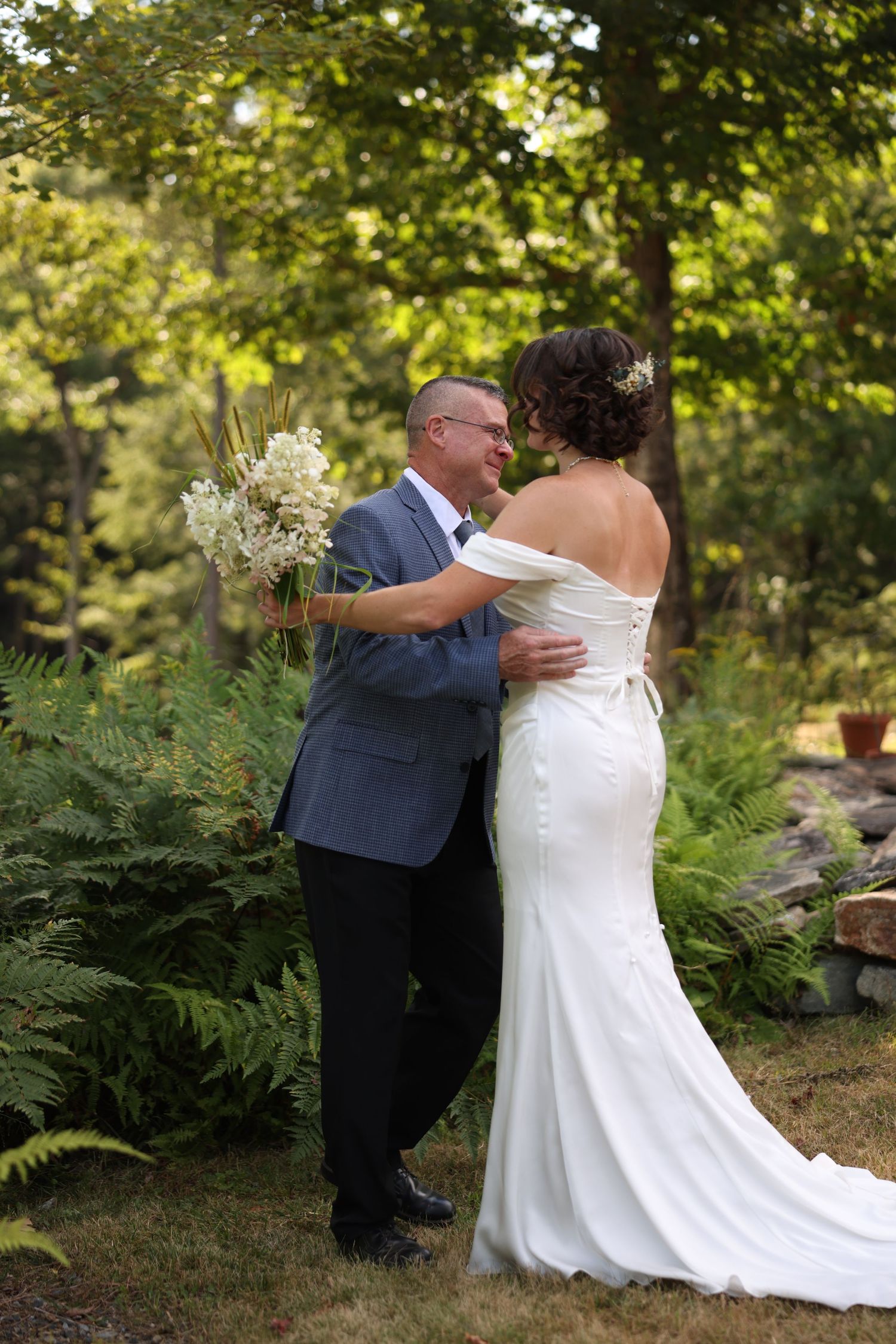 Groom smiles joyfully embracing bride in elegant off-shoulder white gown during first look in garden.