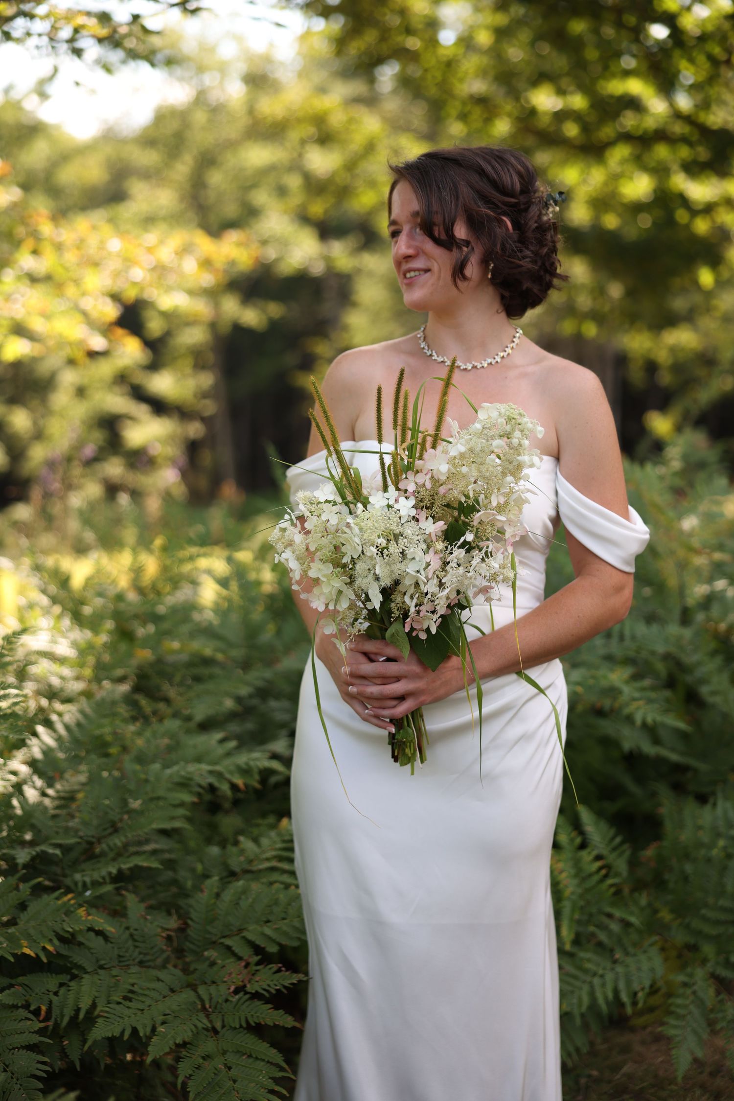 Radiant bride in off-shoulder white gown holds white floral bouquet while smiling in sunlit fern garden.