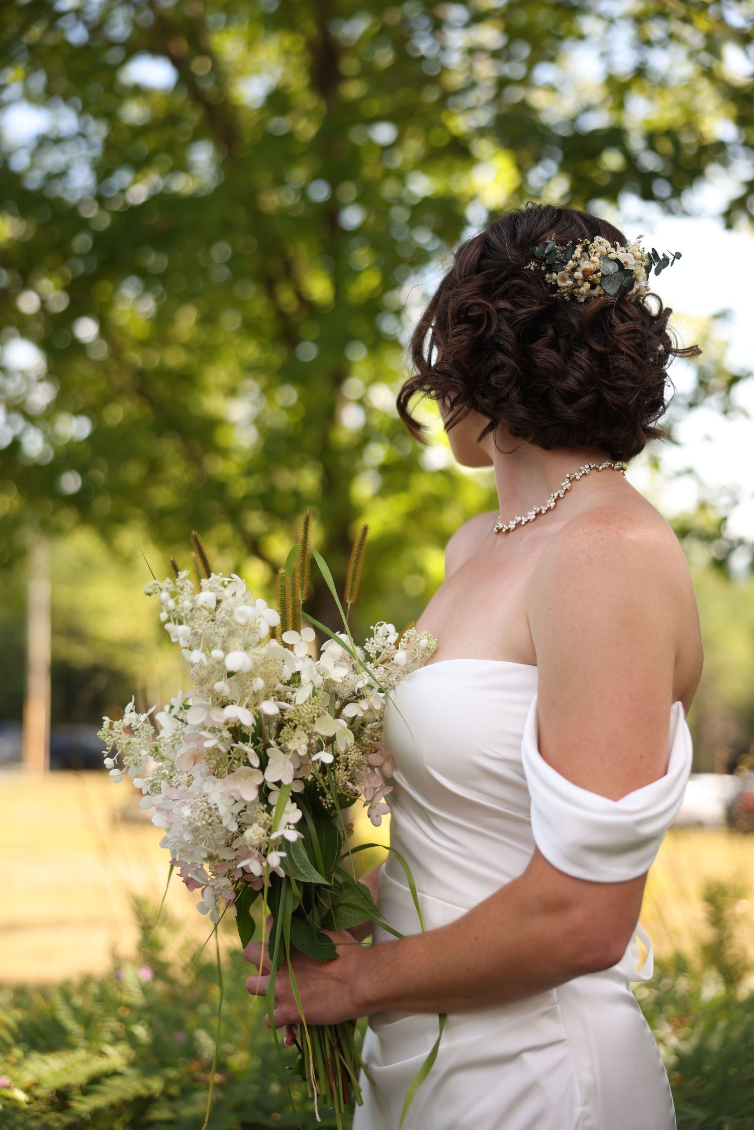 Bride in white off-shoulder gown holding white floral bouquet with updo hairstyle outdoors.