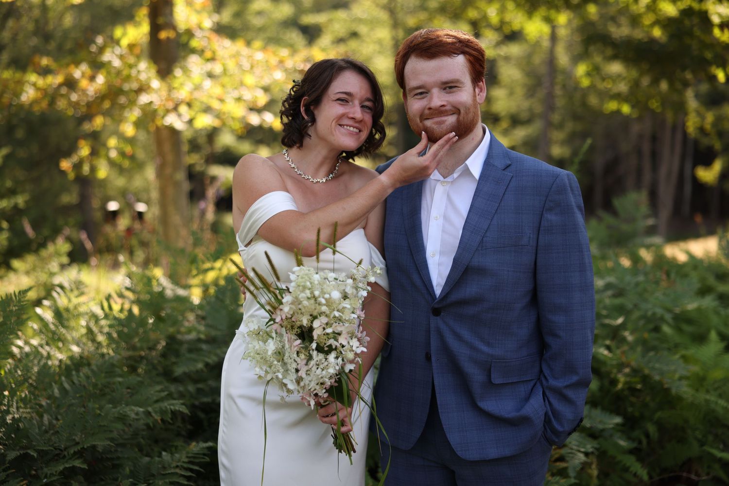 Bride playfully touching grooms chin while holding white bouquet in forest wedding photo.