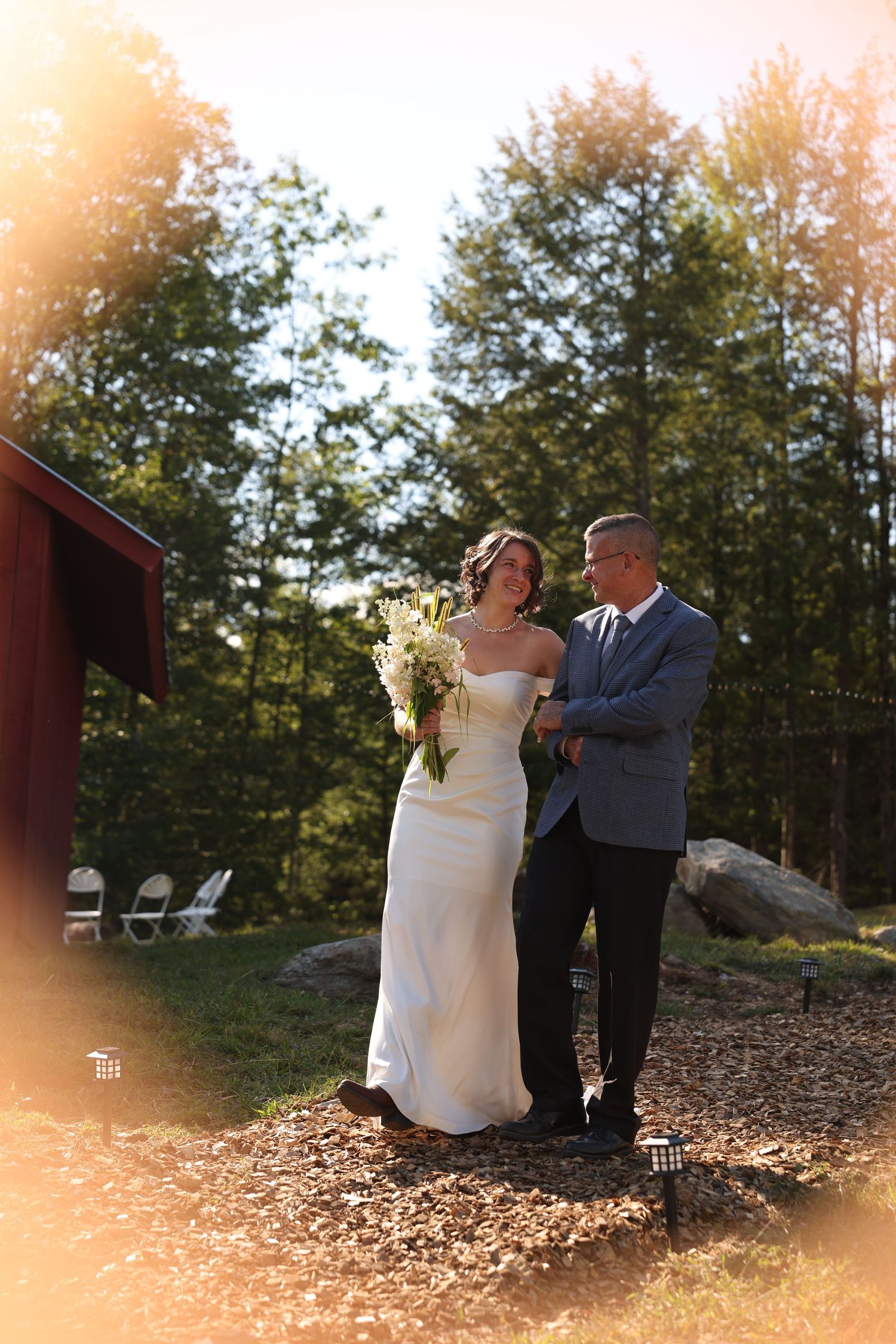 Bride and older gentleman walking together in golden sunlight near red barn at wedding.