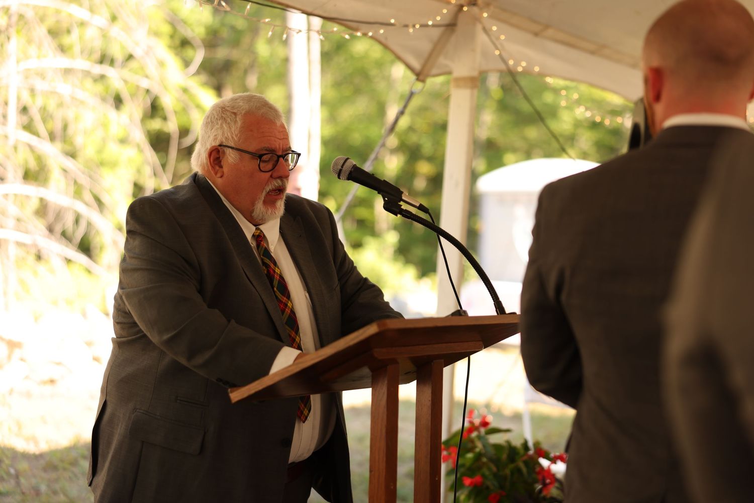 Elderly officiant reading from book at podium during outdoor wedding ceremony under tent.