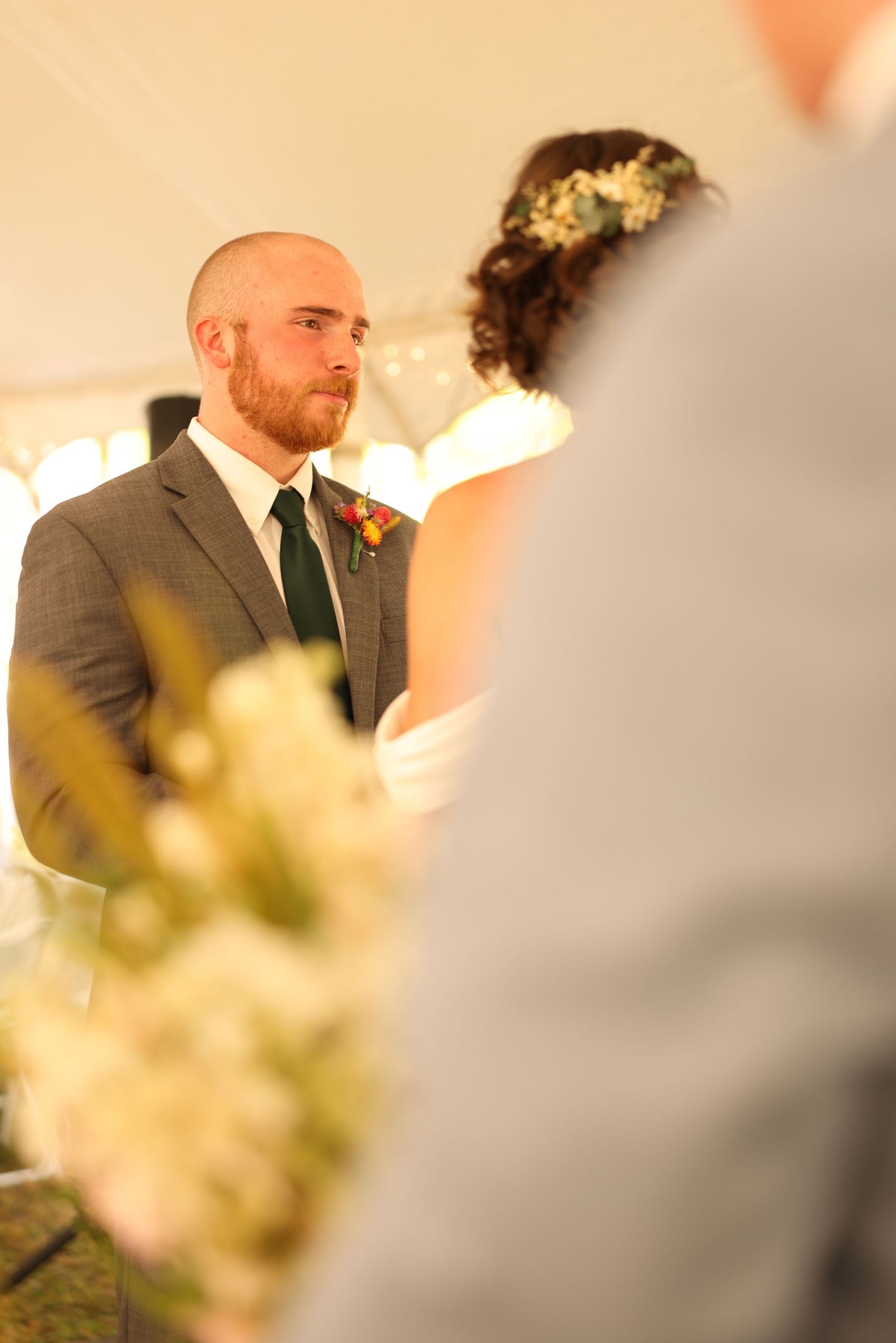 Groom in gray suit with boutonniere gazes lovingly at bride during outdoor wedding ceremony under white tent.