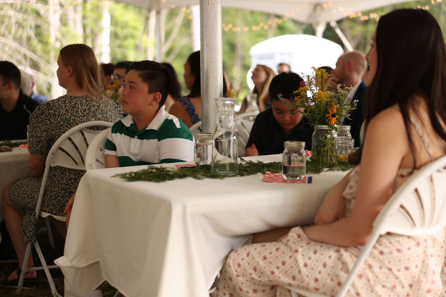 Wedding guests including a young boy in green polo seated at white linen tables under an elegant outdoor tent.