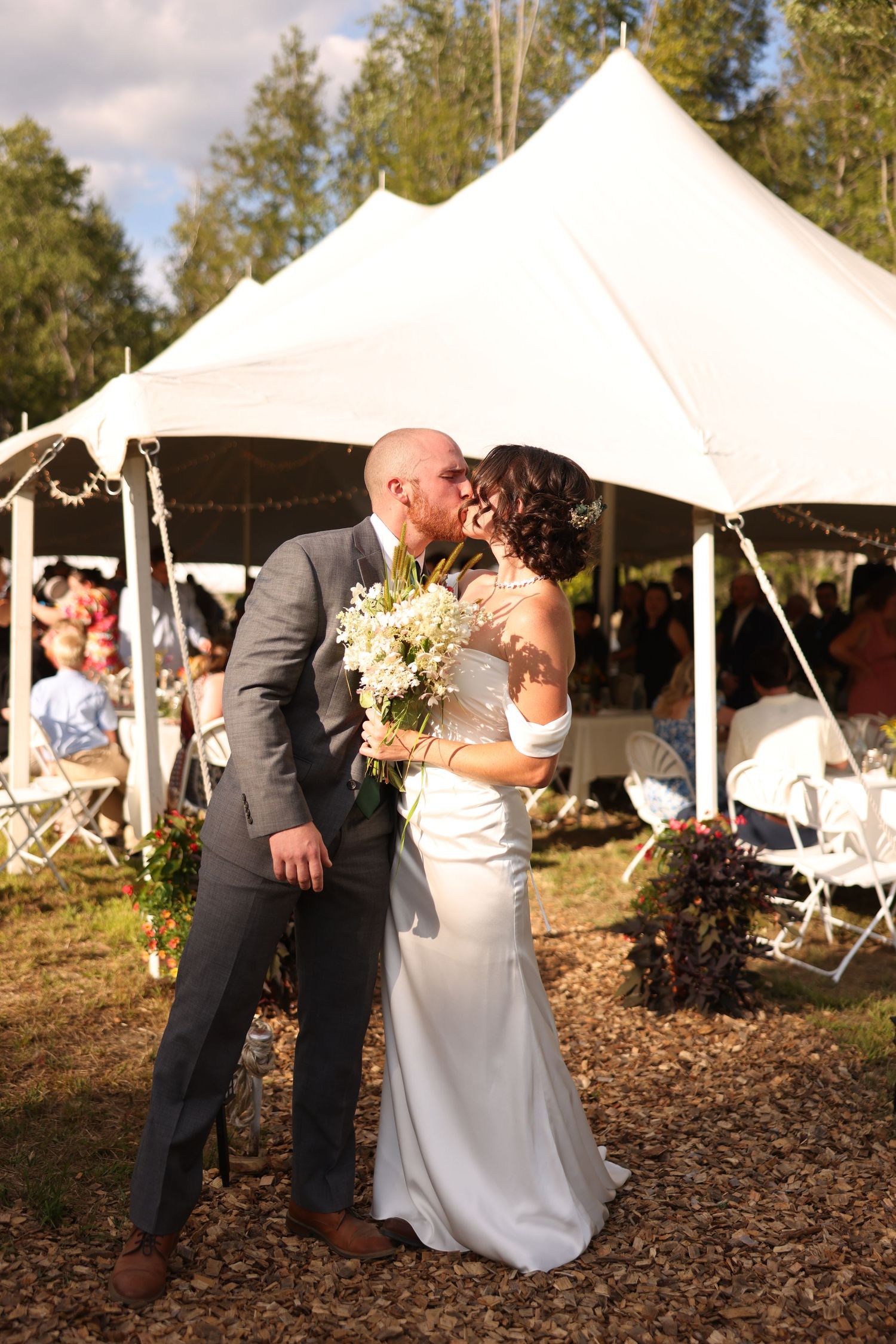 Newlyweds share first kiss outside white wedding tent surrounded by trees and seated guests.