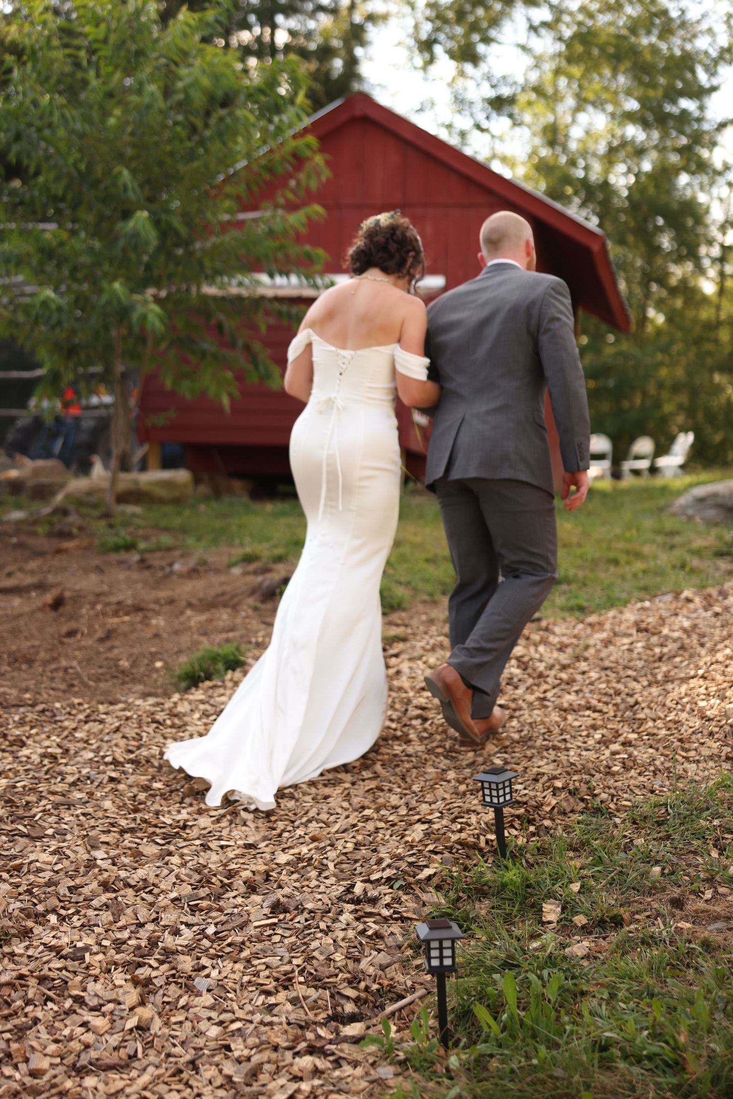 Bride in fitted white off-shoulder gown and groom in gray suit walk toward red barn on wood chip path.
