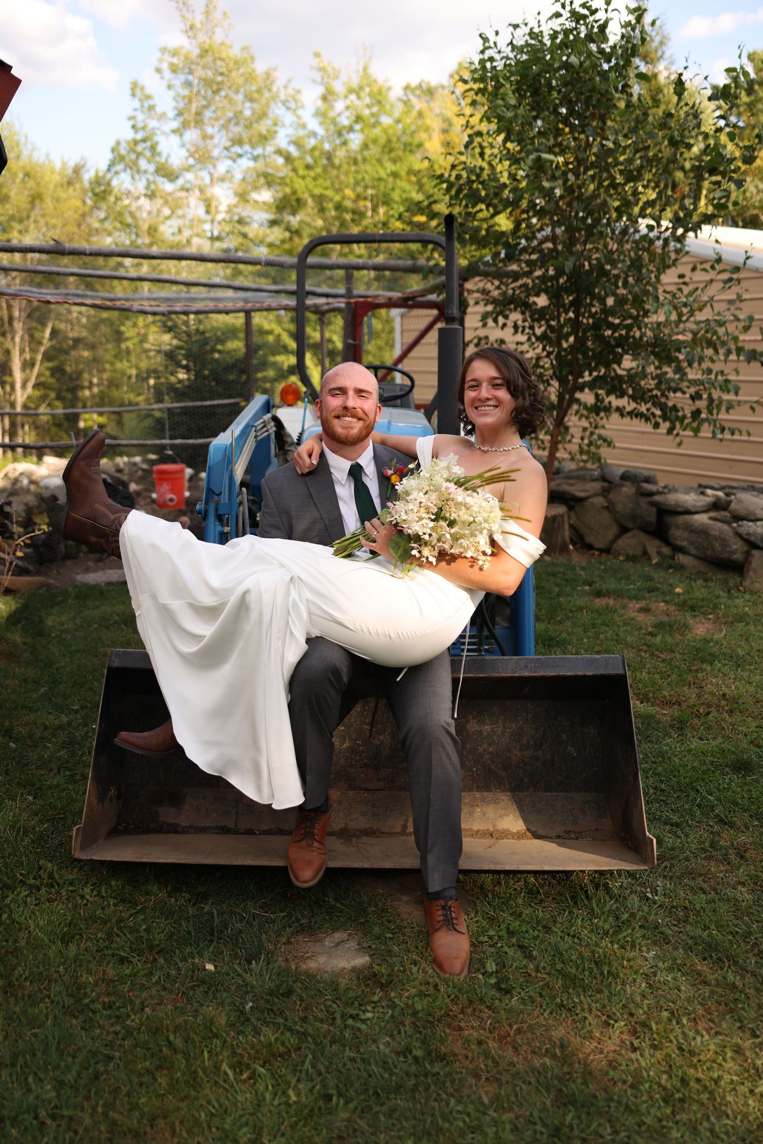 Groom holds bride in white gown while seated in tractor bucket loader at rustic outdoor wedding venue.