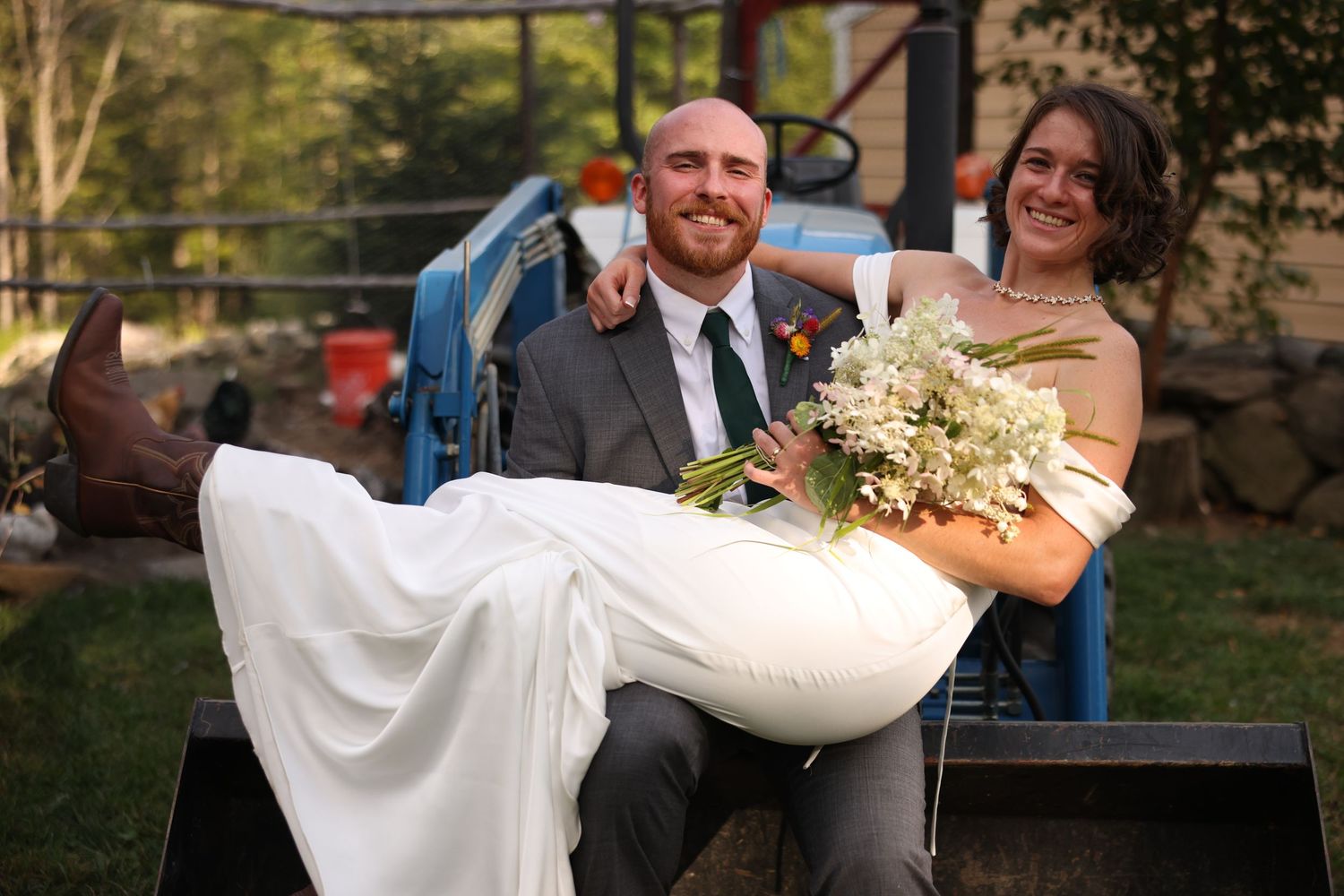 Smiling bride and groom pose playfully on blue farm tractor holding white floral bouquet at rustic wedding.