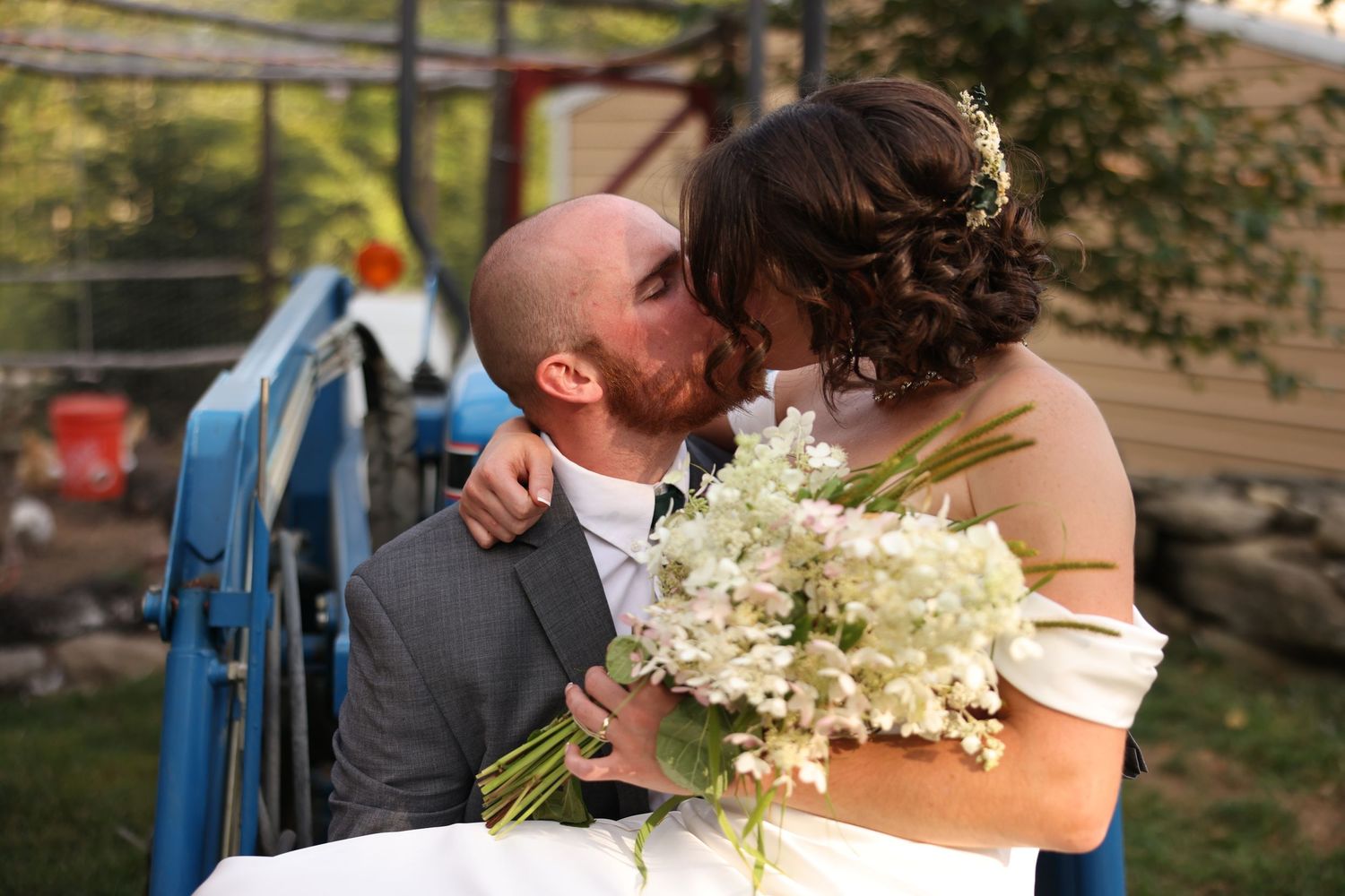 Bride and groom share romantic kiss near blue tractor as bride holds white floral bouquet at farm wedding.