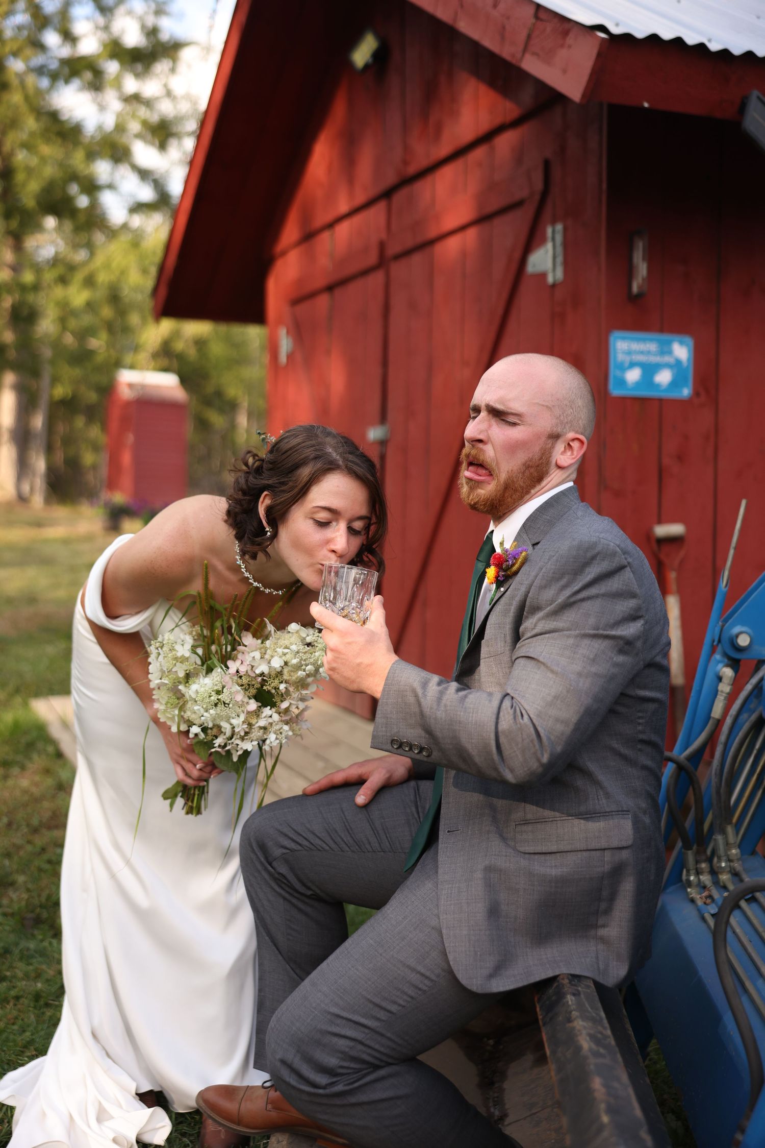 Bride kisses grooms hand while he makes funny face seated by red barn at rustic farm wedding.