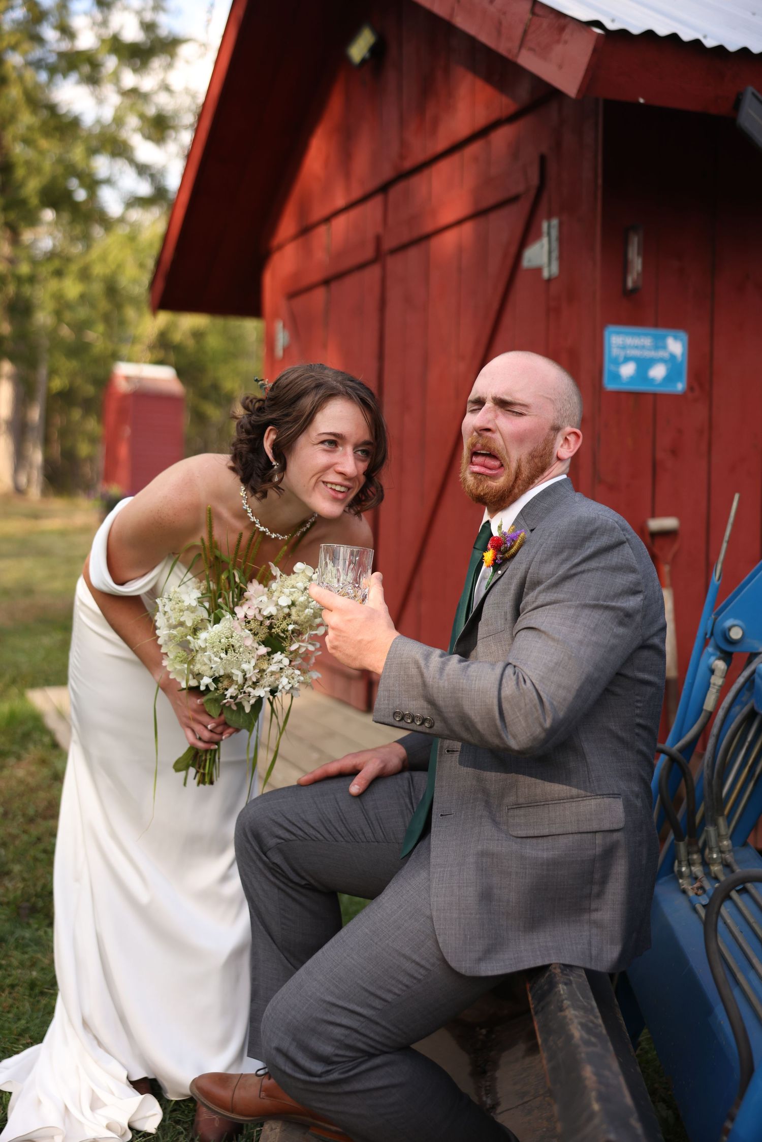 Bride laughs while groom makes humorous expression holding drink glass beside red barn at farm wedding.