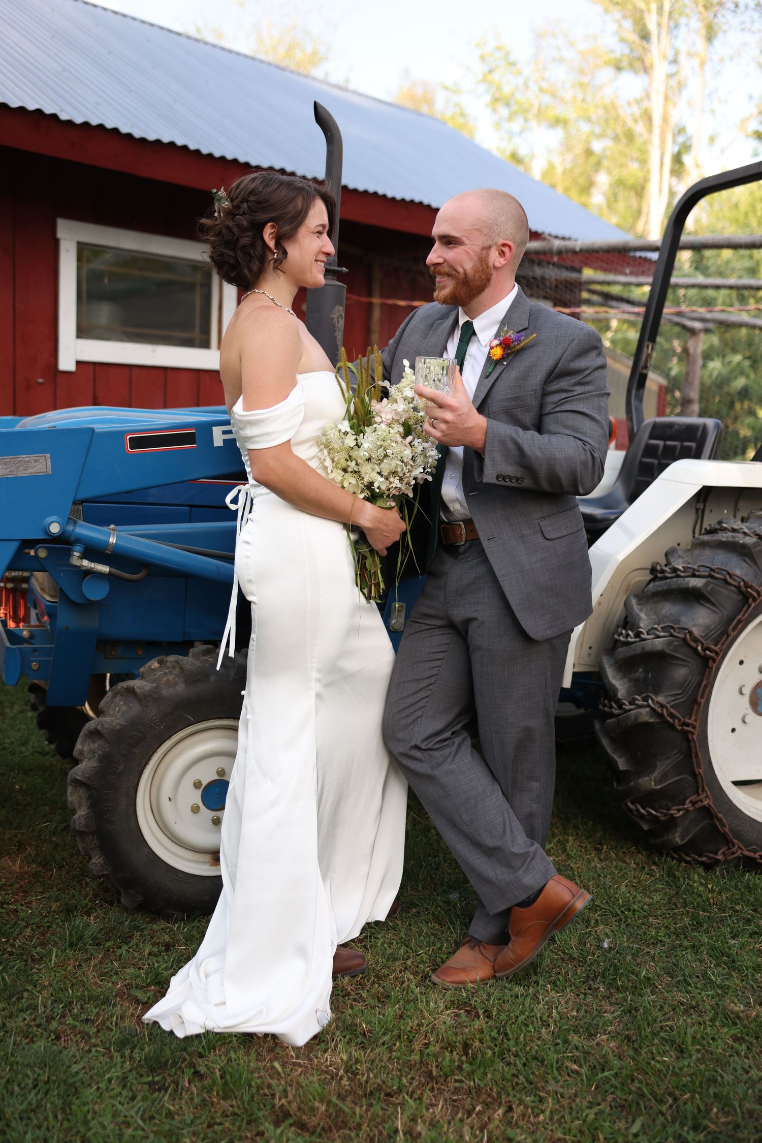 Bride and groom smile at each other holding bouquet and drink beside vintage blue tractor at farm wedding.