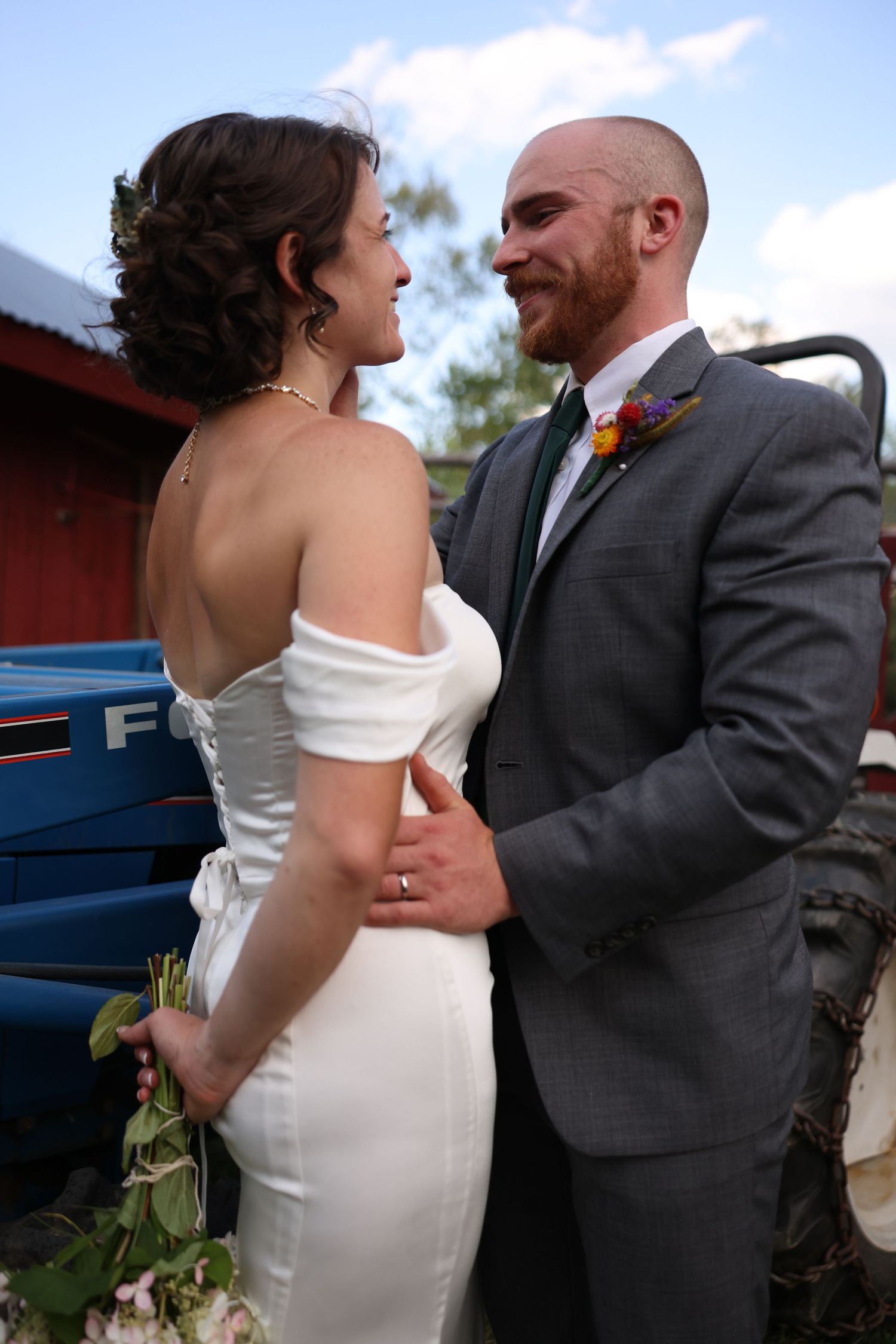 Smiling bride and groom embrace near a blue tractor at a rustic farm wedding venue.