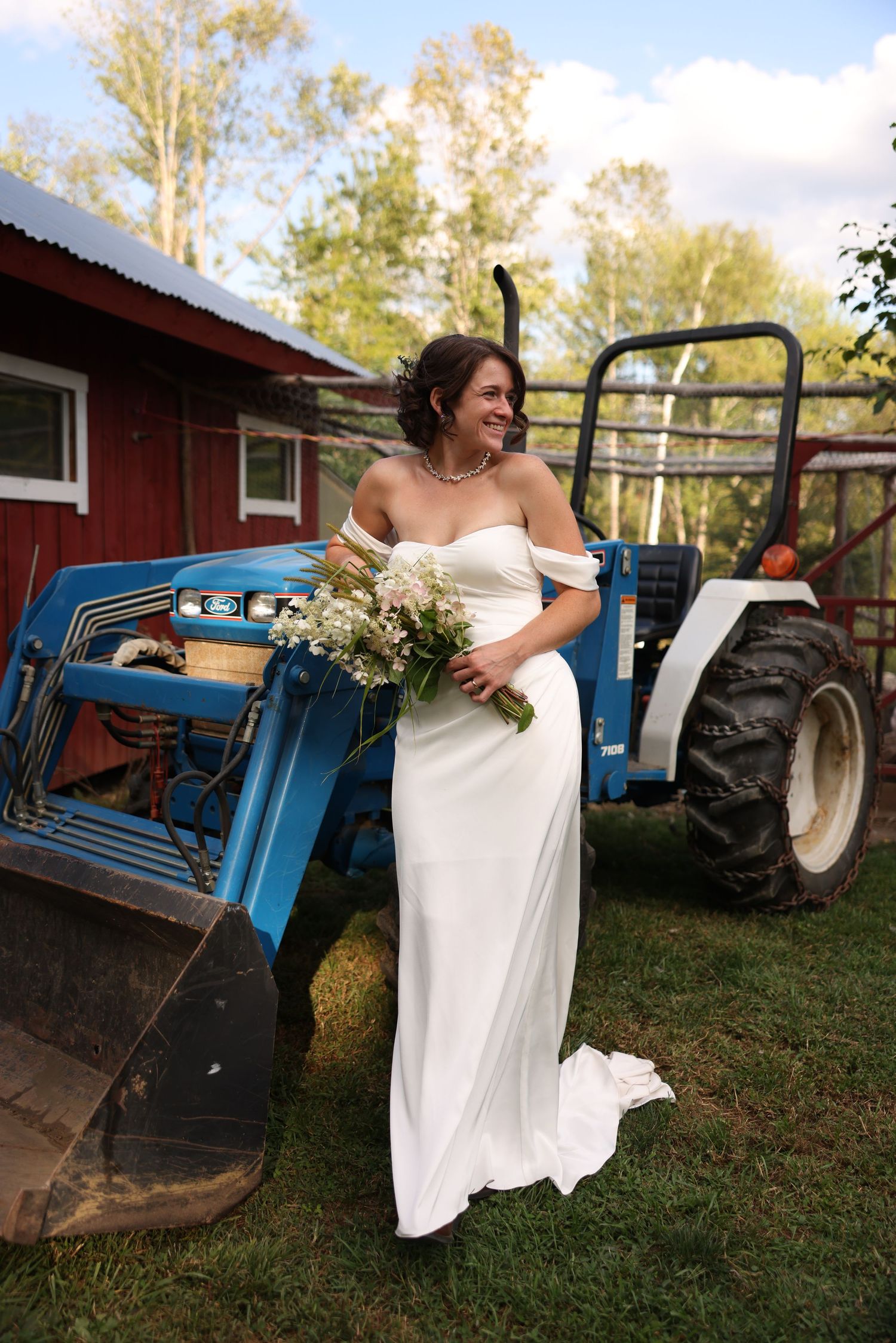 Bride in off-shoulder white gown holds wildflower bouquet beside a blue tractor at a farm.