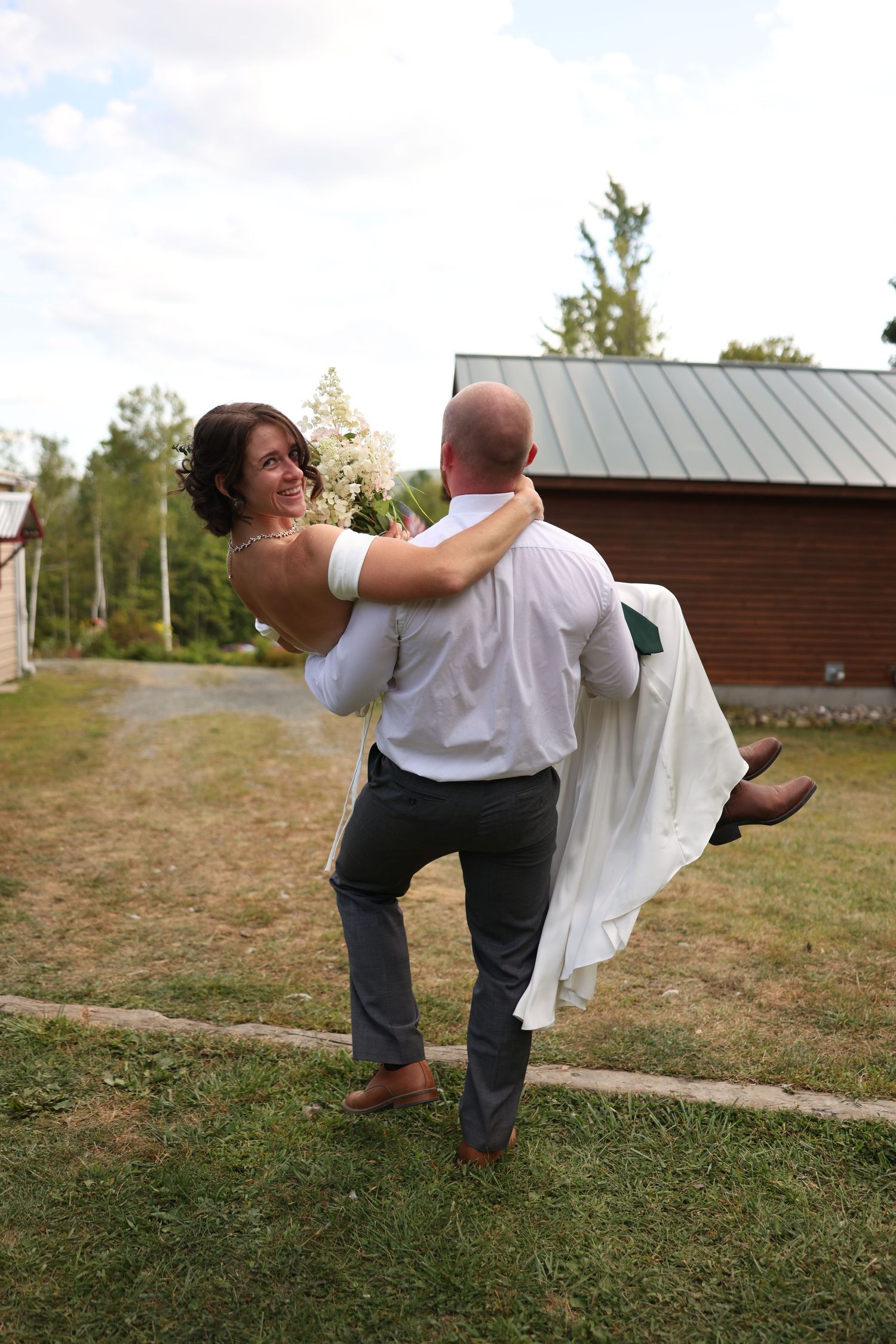 Groom lifts smiling bride in white dress outdoors near a rustic barn on their wedding day.