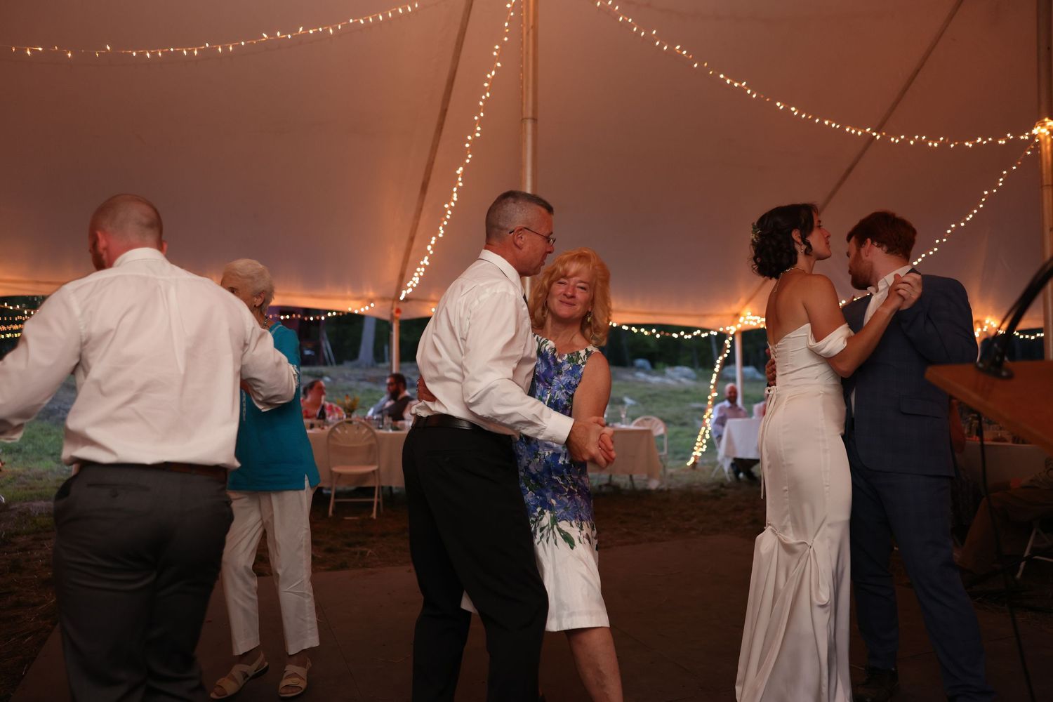 Guests and newlyweds dance joyfully together under glowing string lights at wedding reception.