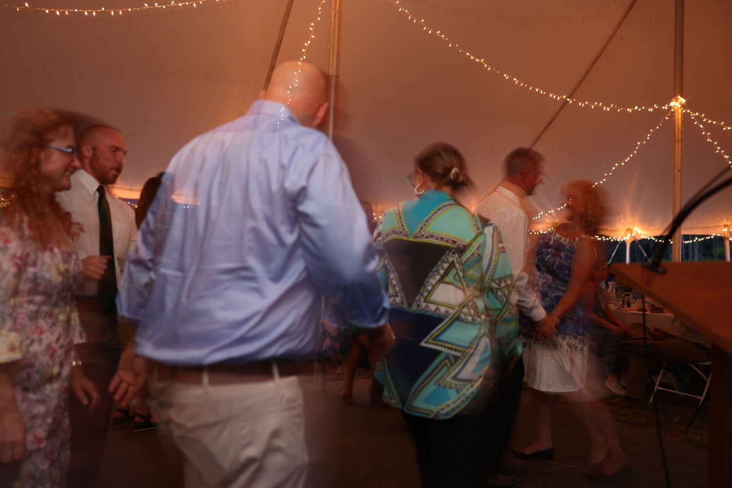 Wedding guests dance together under a warmly lit tent at an evening outdoor reception.