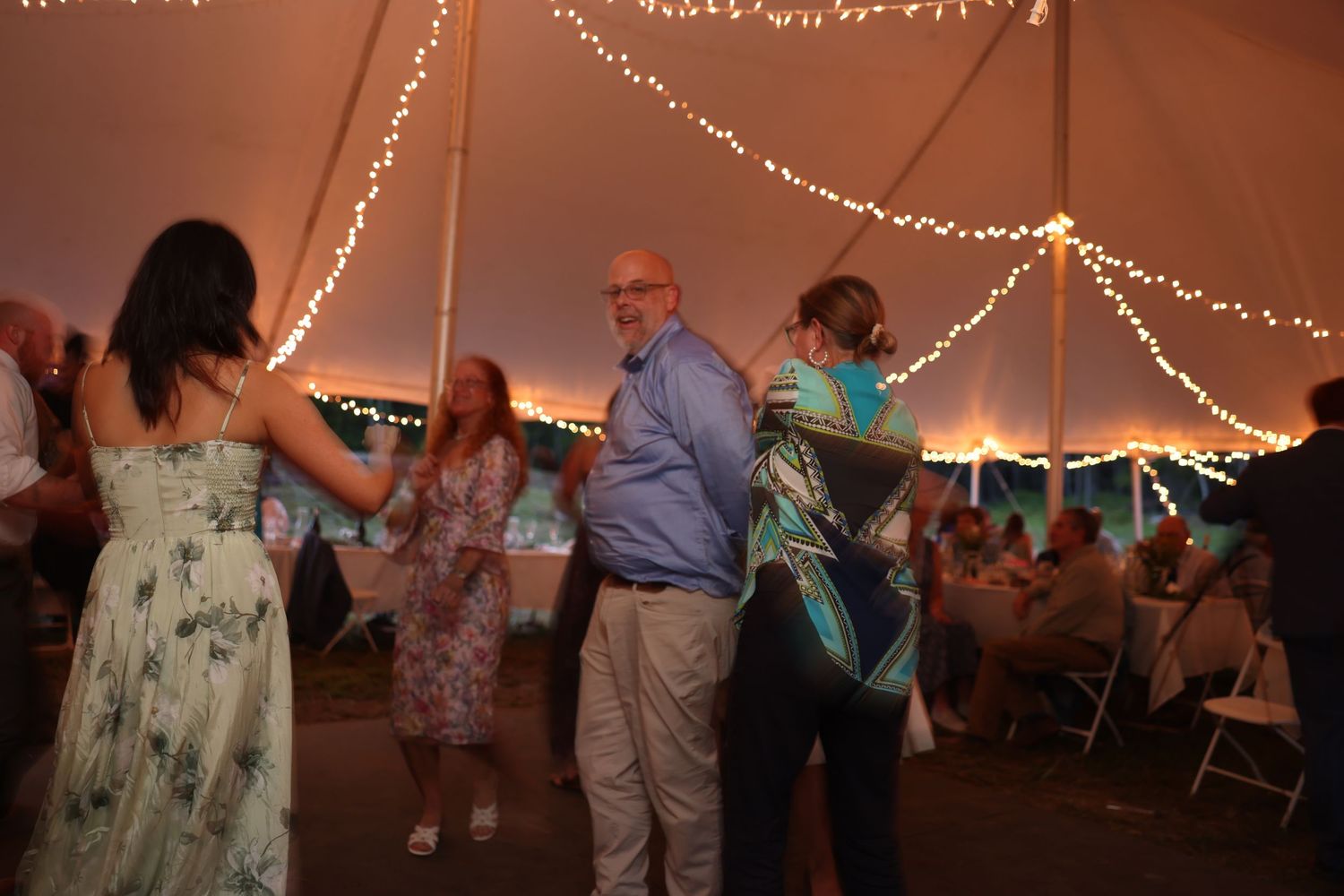 Guests dancing under a warmly lit tent with string lights at an elegant outdoor wedding reception.