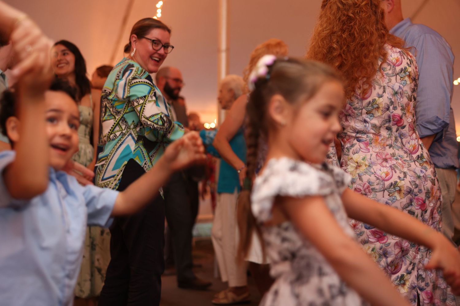 Children and adults joyfully dancing together inside a festively lit wedding reception tent.