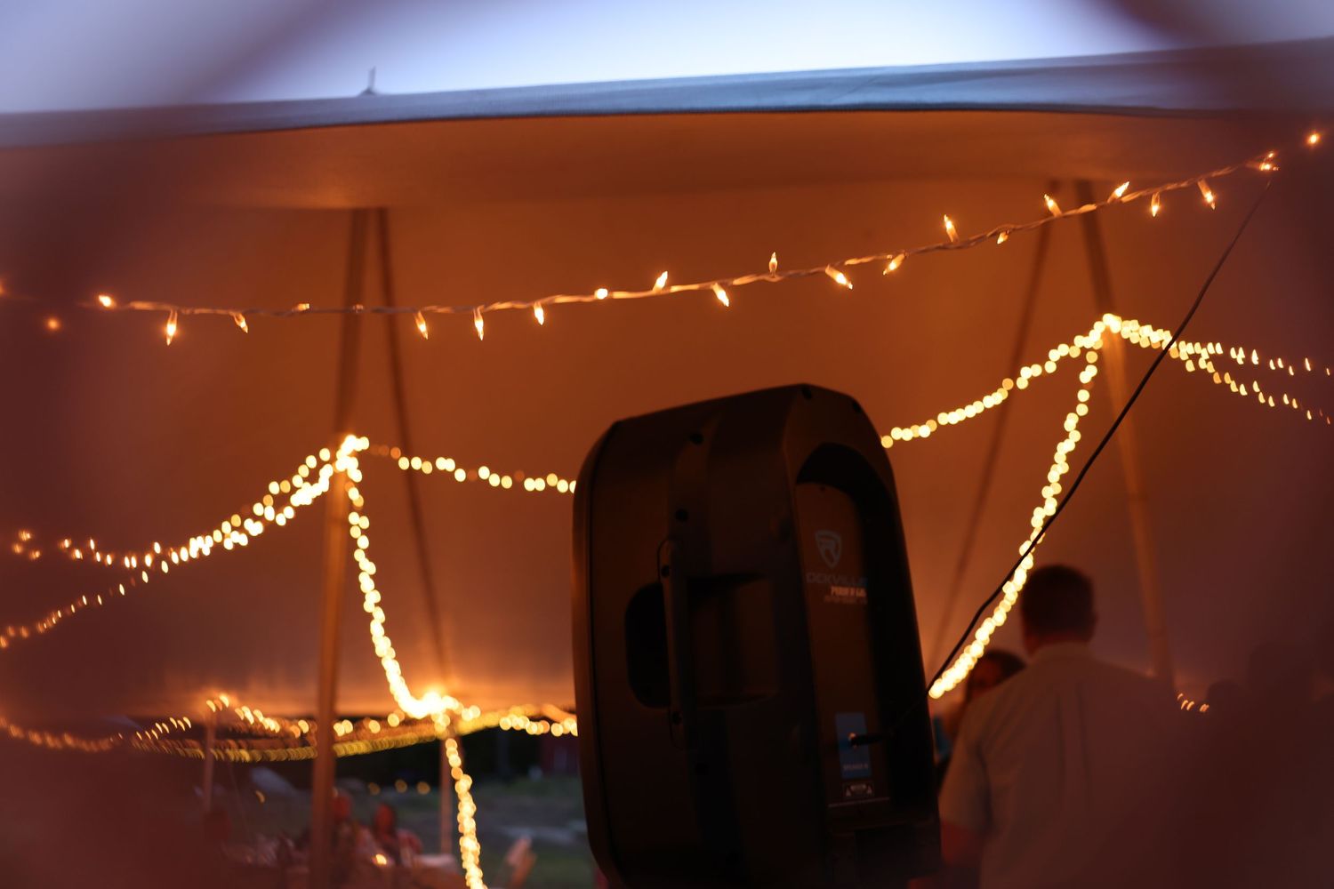 Warm golden string lights draped across a wedding tent ceiling with a speaker in the foreground.