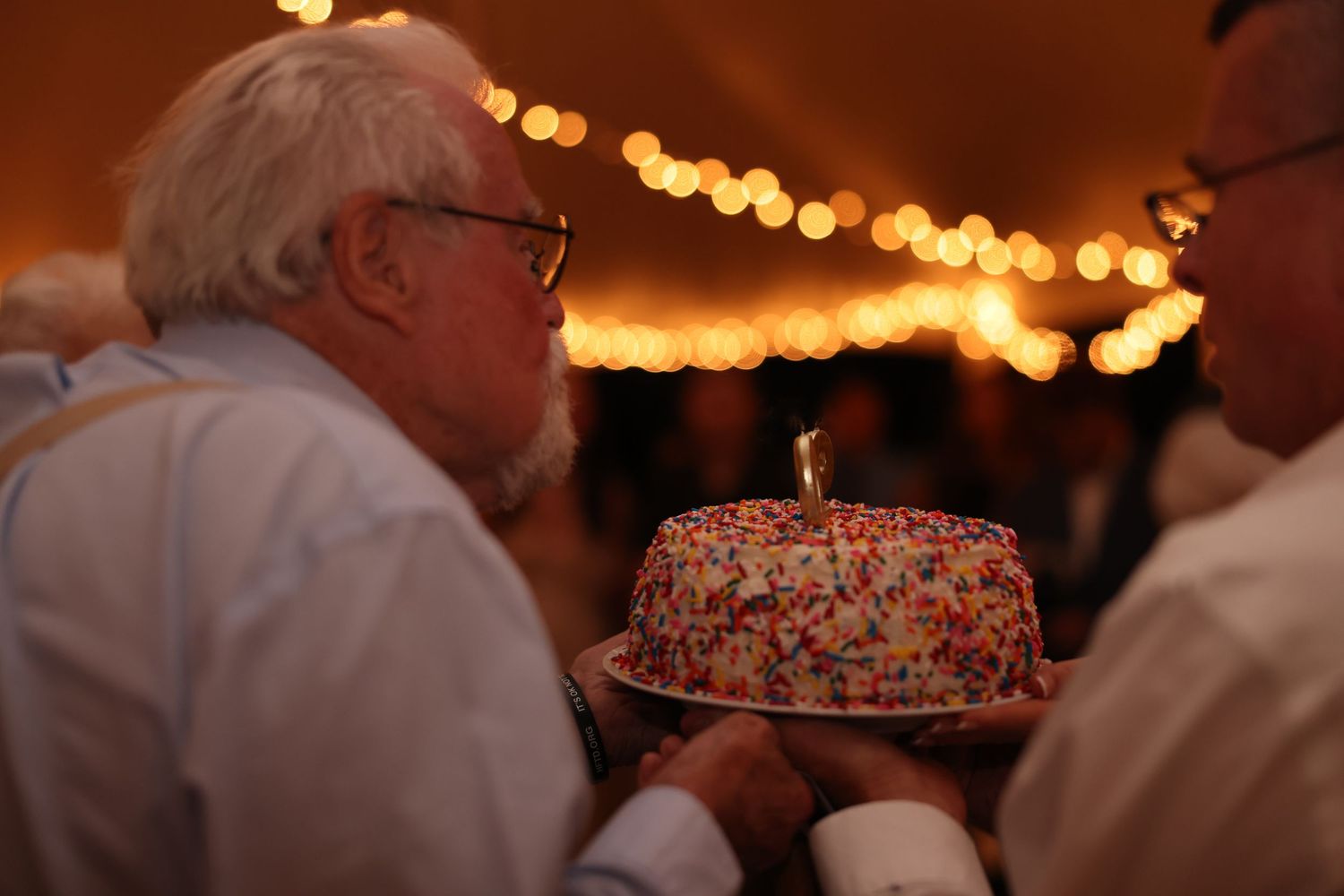 Elderly man leaning in to blow out candles on a colorful sprinkle birthday cake at a party.