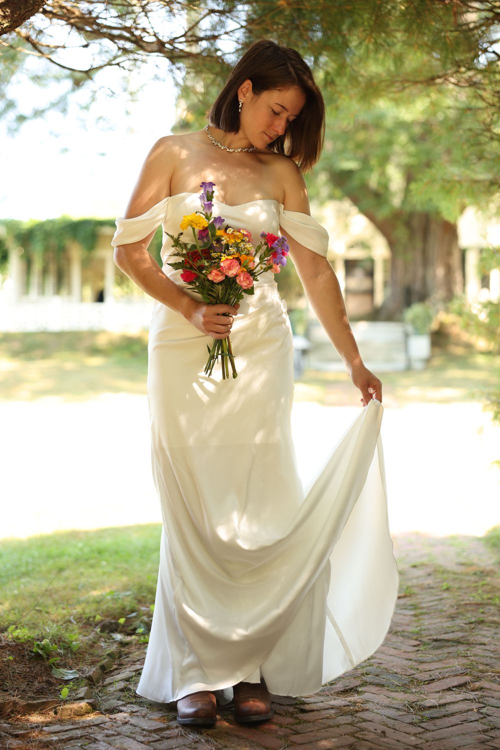Bride in off-shoulder satin wedding gown holding colorful bouquet while lifting dress on cobblestone.