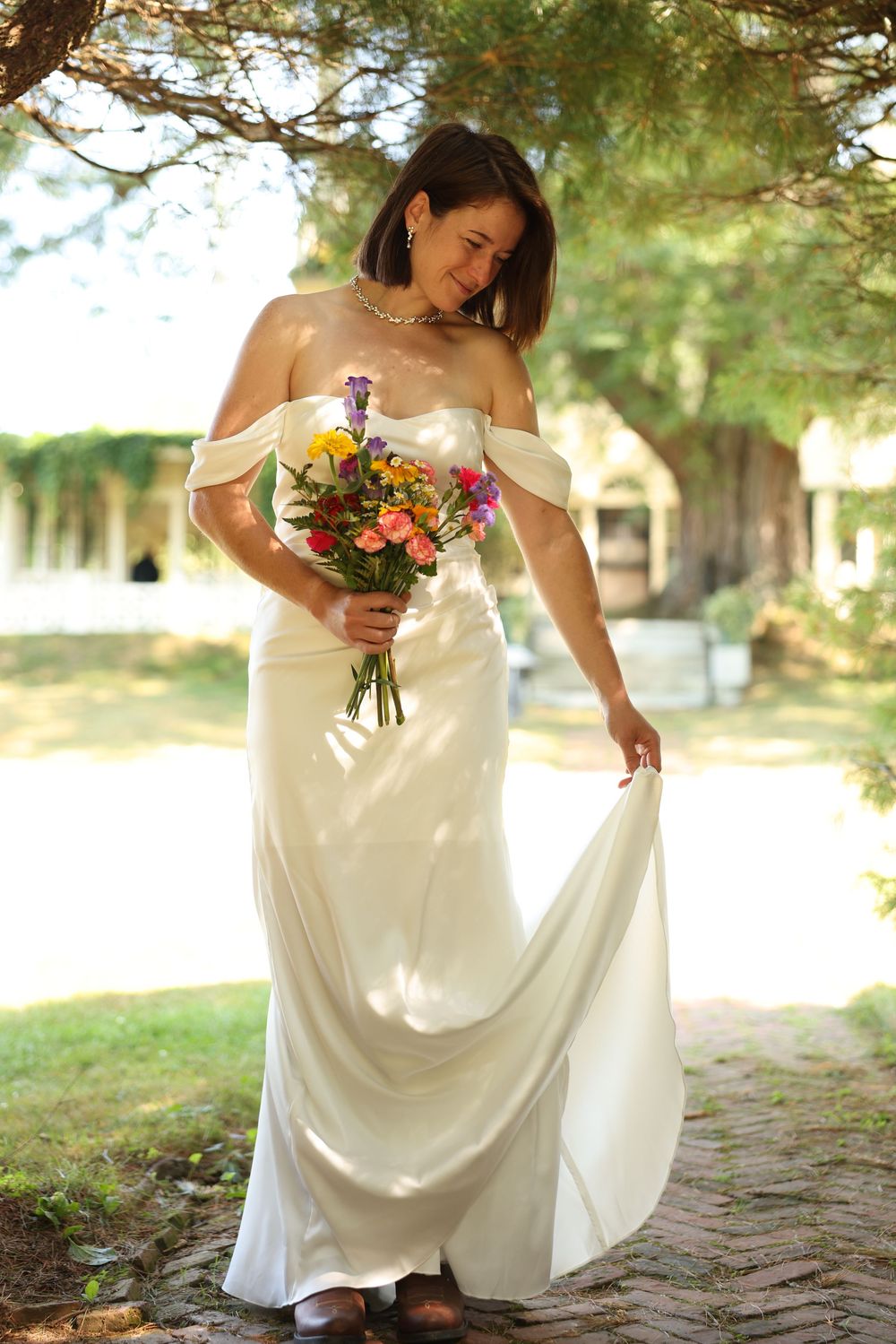 Elegant bride in off-shoulder satin gown holding colorful bouquet lifting dress hem under trees.