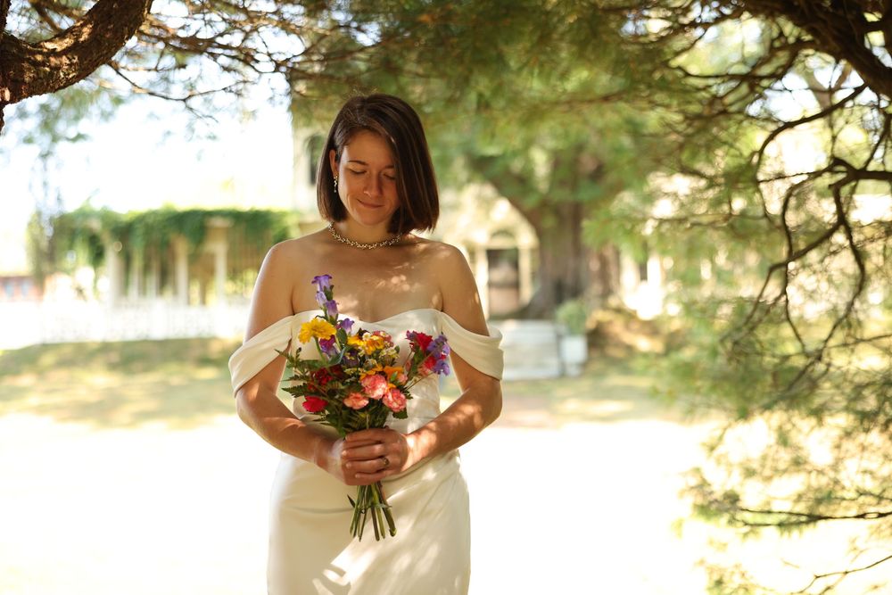 Smiling bride in off-shoulder white gown holding colorful wildflower bouquet outdoors under trees.