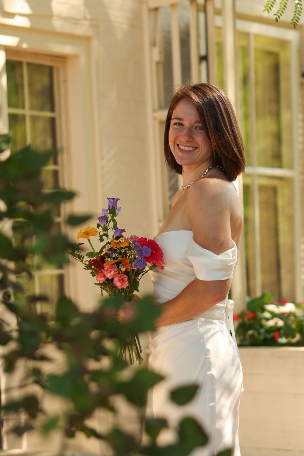 Happy bride in strapless white gown holding vibrant bouquet smiling outside elegant white building.