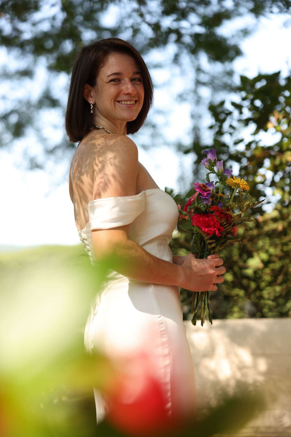 Radiant bride in off-shoulder white gown smiling while holding colorful floral bouquet outdoors.