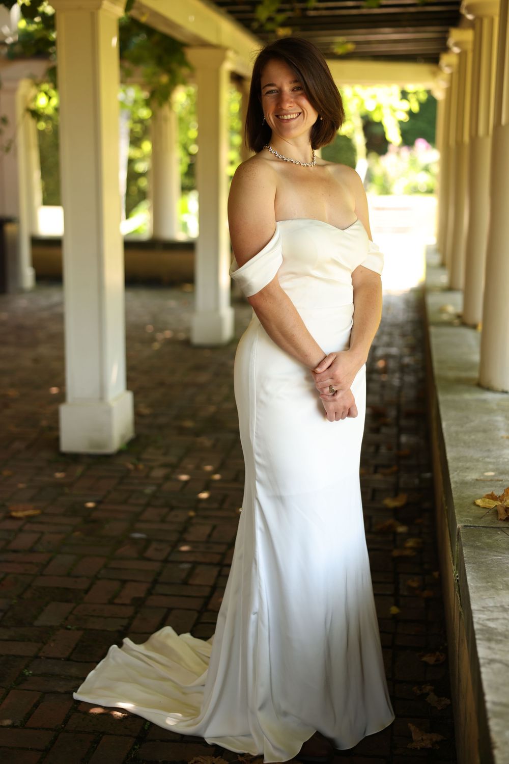 Smiling bride in elegant off-shoulder white gown standing in a sunlit garden colonnade.