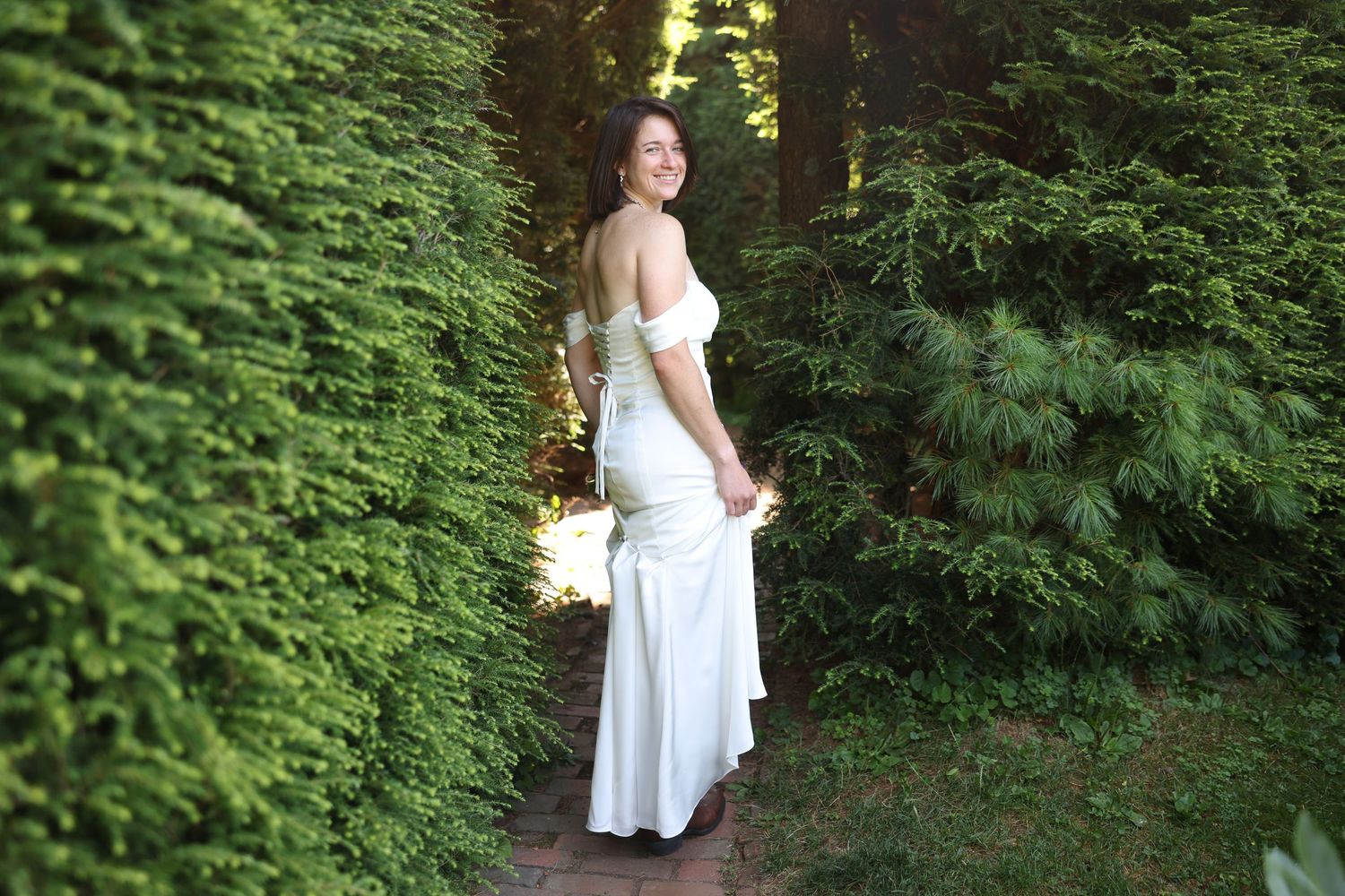 Bride in white off-shoulder wedding gown walking along a lush green garden pathway.